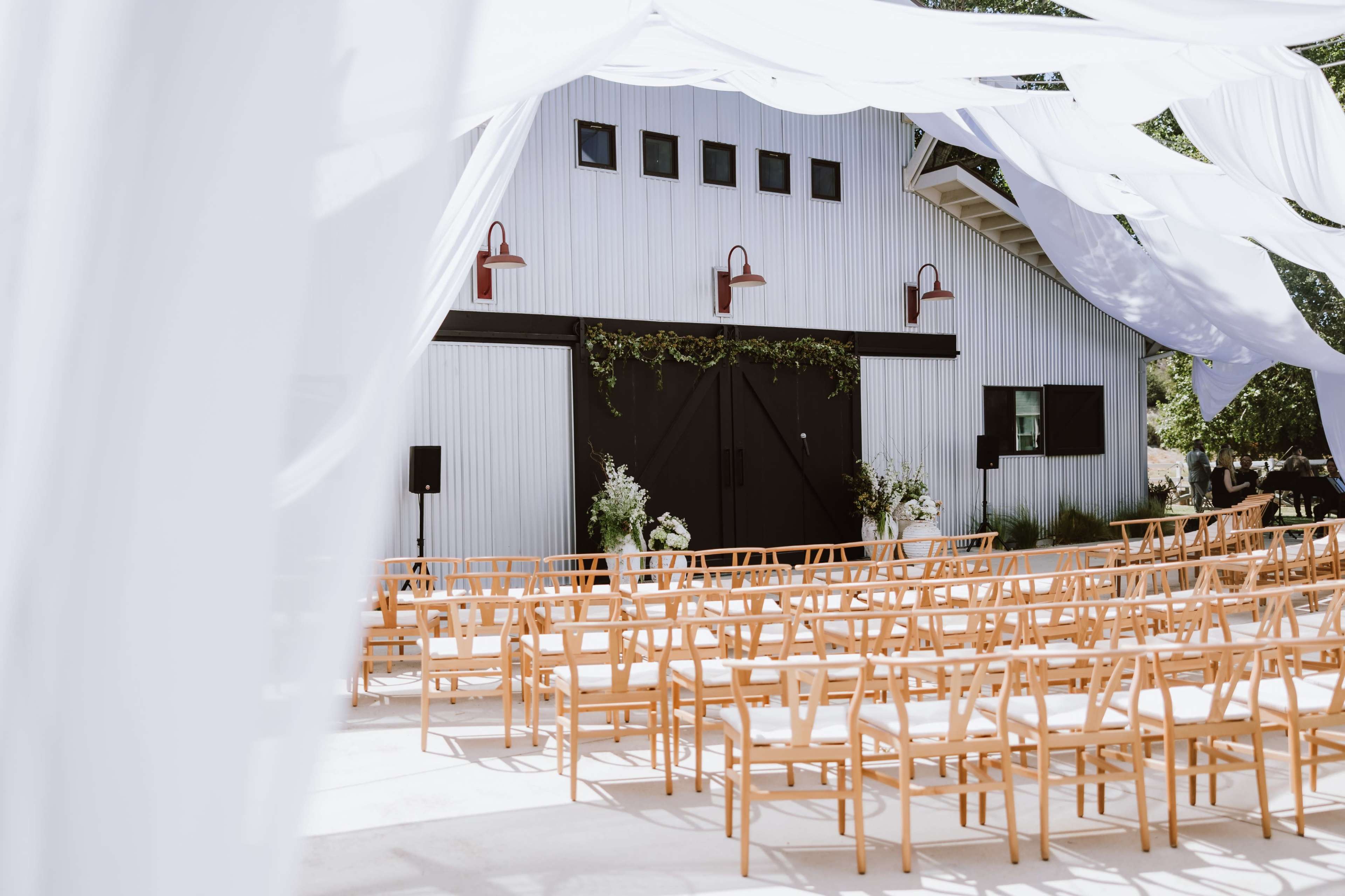 A wedding venue is set up outdoors with rows of wooden chairs facing a decorated barn entrance draped in white fabric.