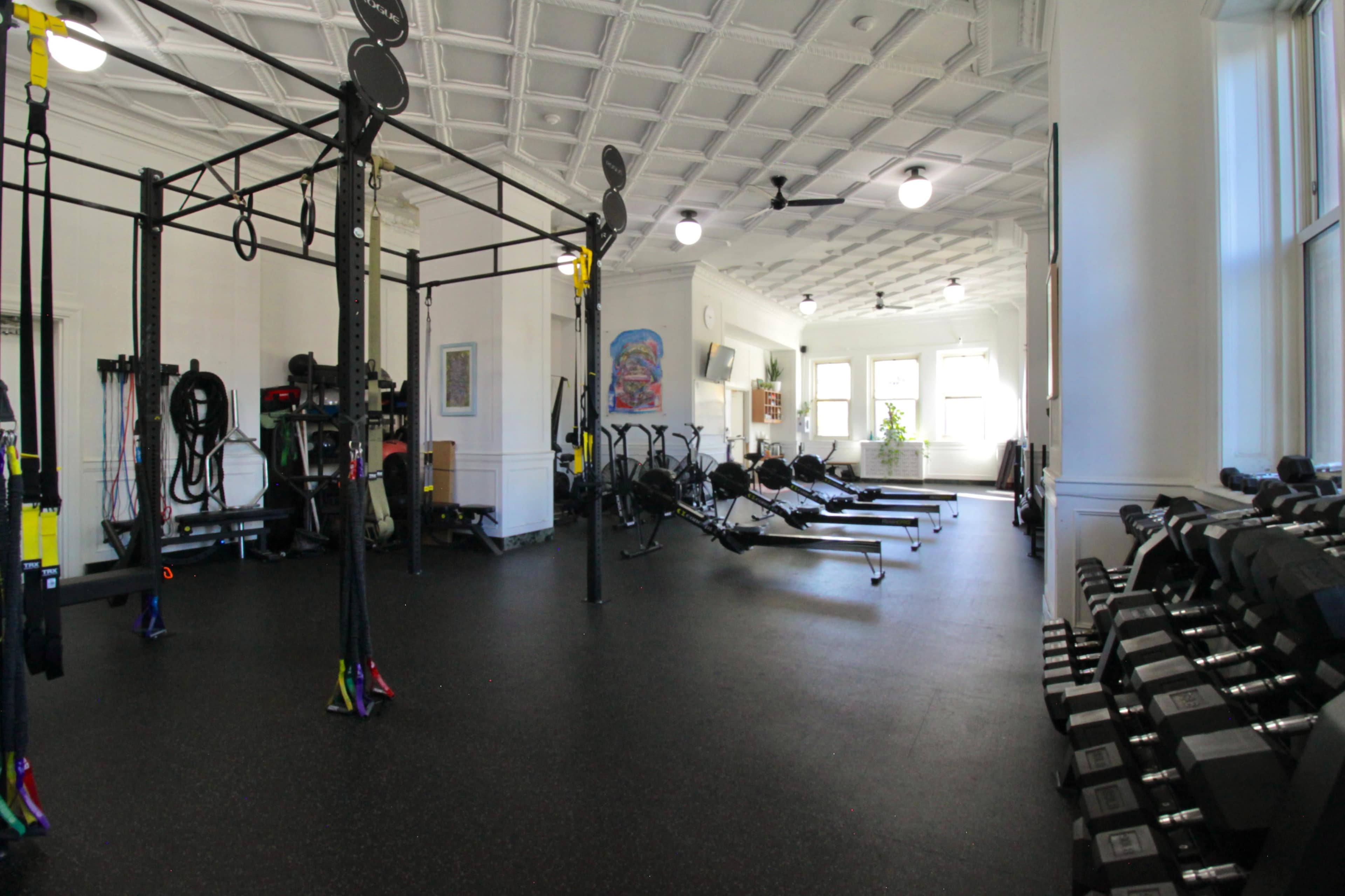 The image shows a spacious gym with various exercise equipment, including rowing machines, weights, and a functional training rig.