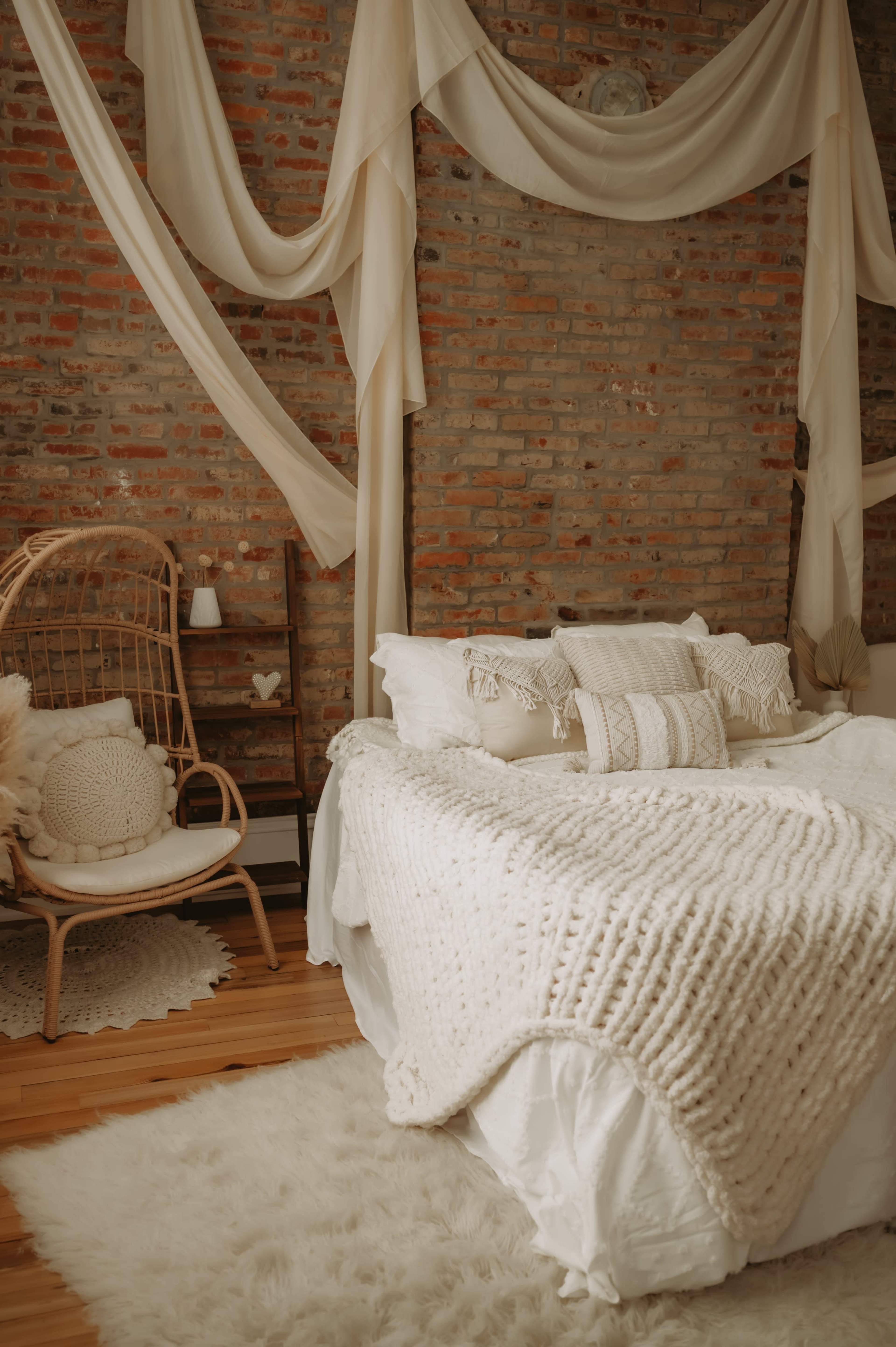 The image shows a cozy bedroom with a knitted blanket on a bed, a rattan chair with decorative cushions, and draped fabric against a brick wall.