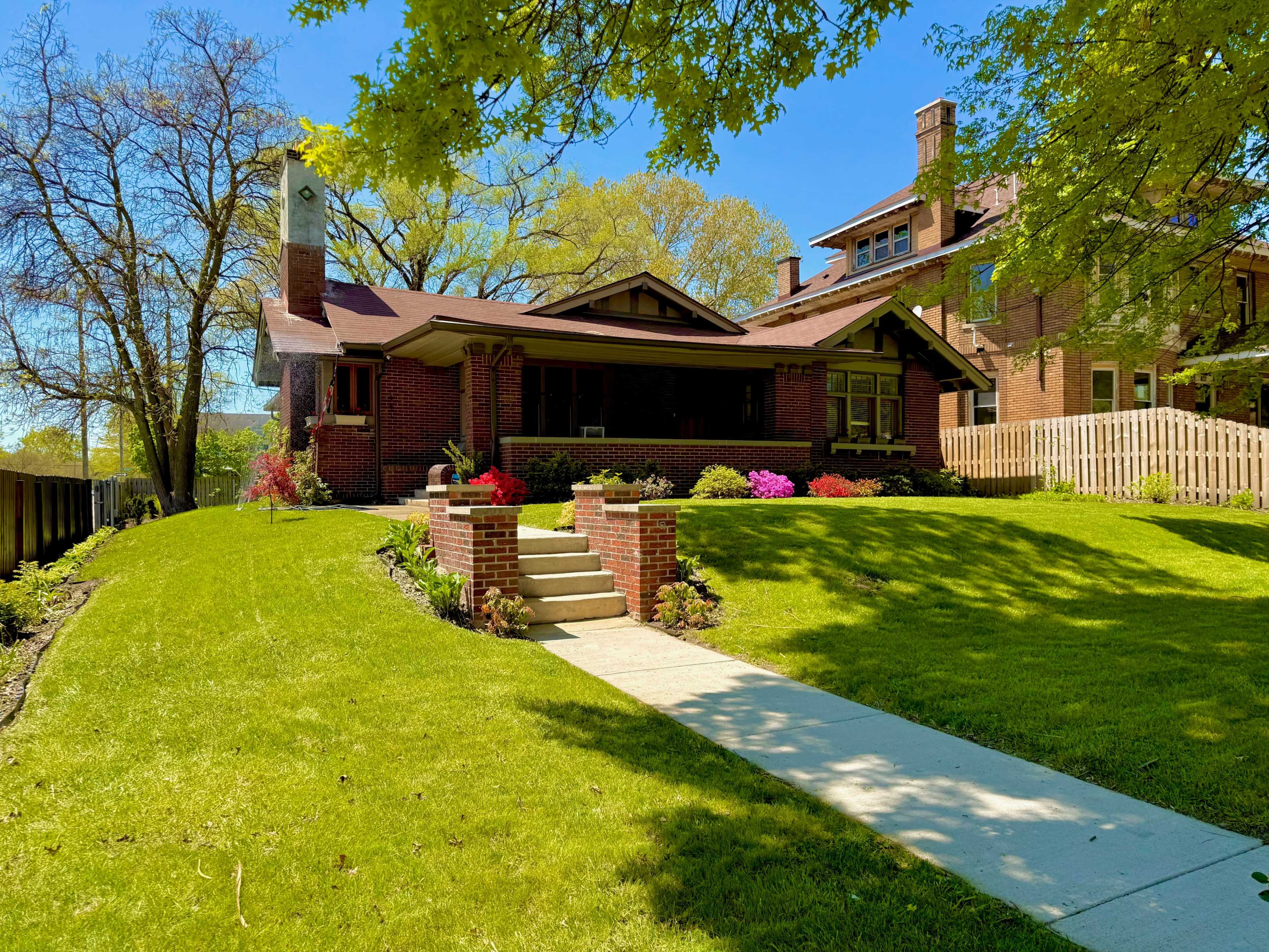 The image shows a well-maintained brick house with a front porch, surrounded by a green lawn and colorful flower beds, under a clear blue sky.