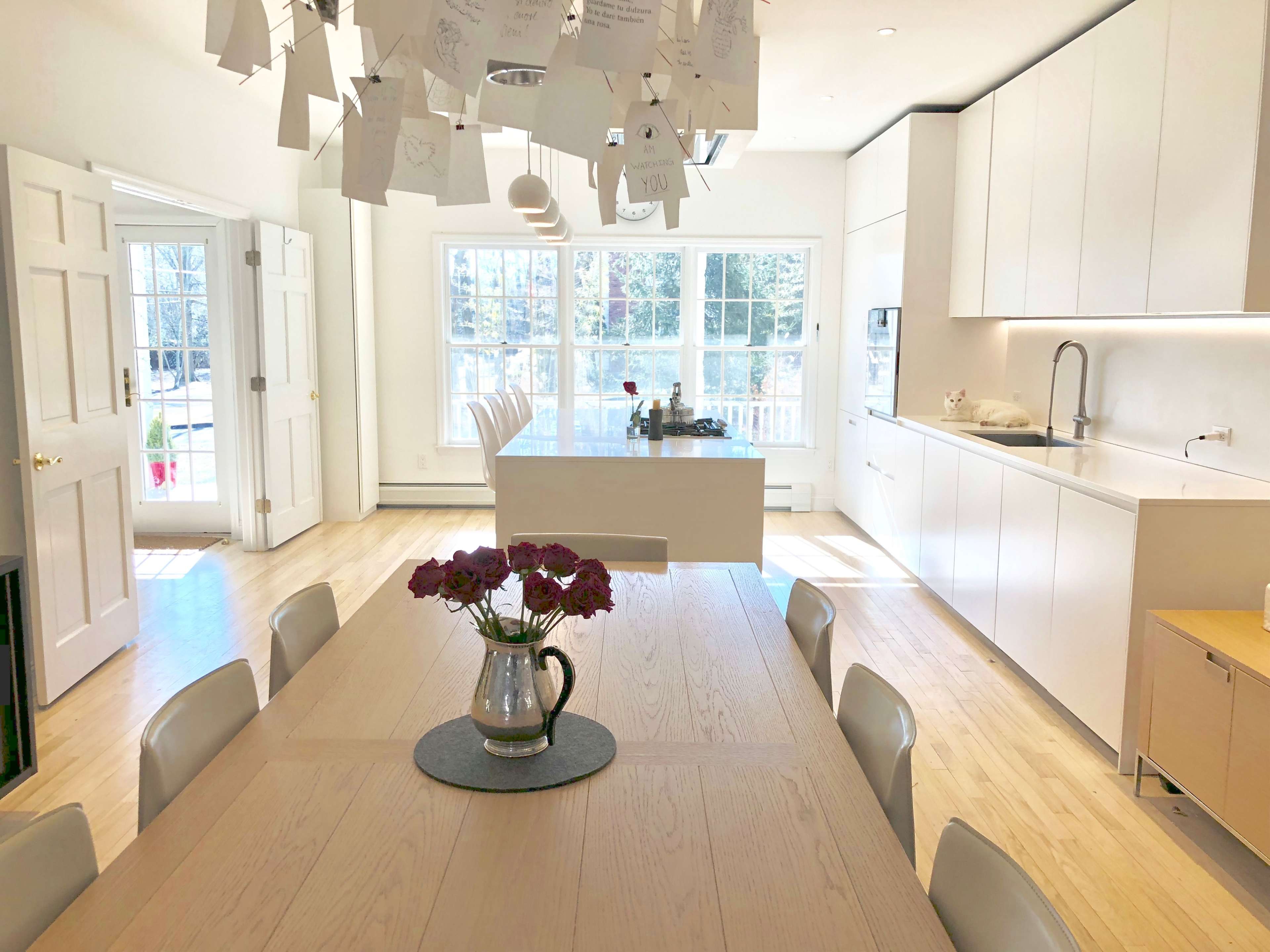 A modern kitchen with a long dining table adorned with a vase of flowers, surrounded by white chairs and bright windows illuminating the space.