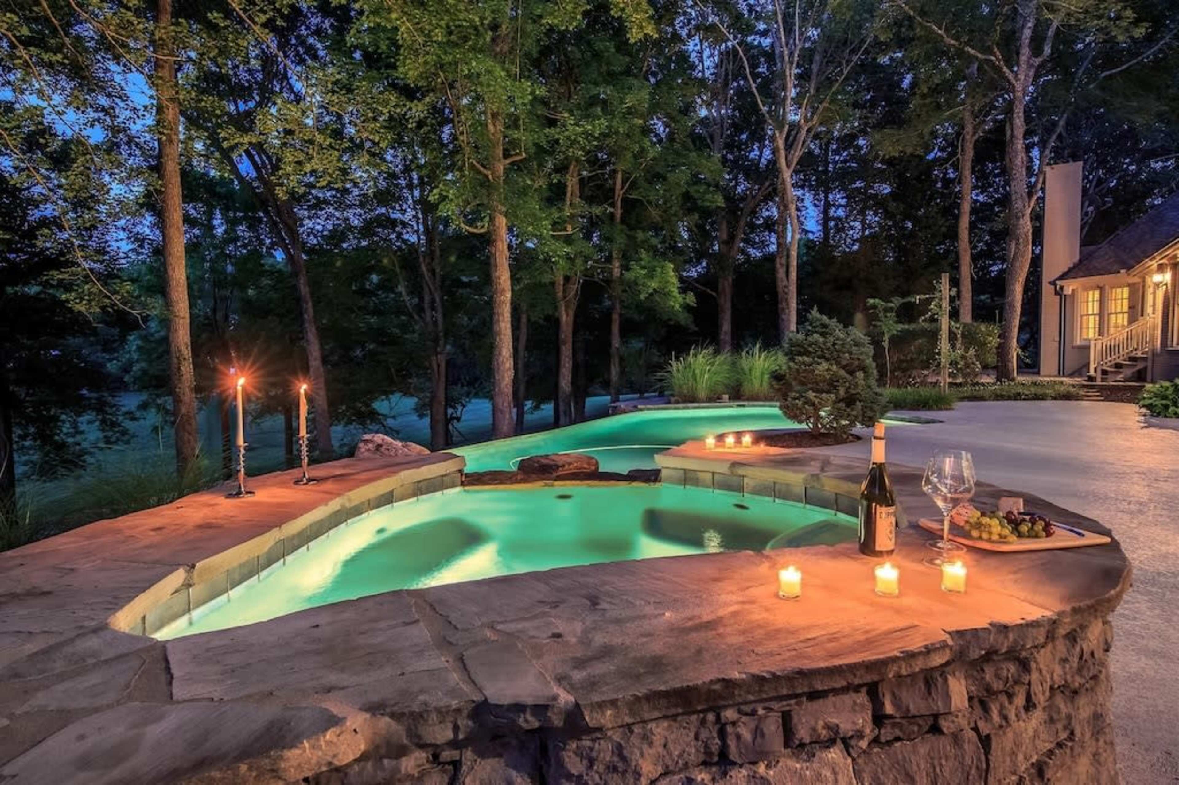 A stone patio features a circular hot tub with glowing water, surrounded by trees and softly lit candles, next to a bottle of wine and a glass on a small table.