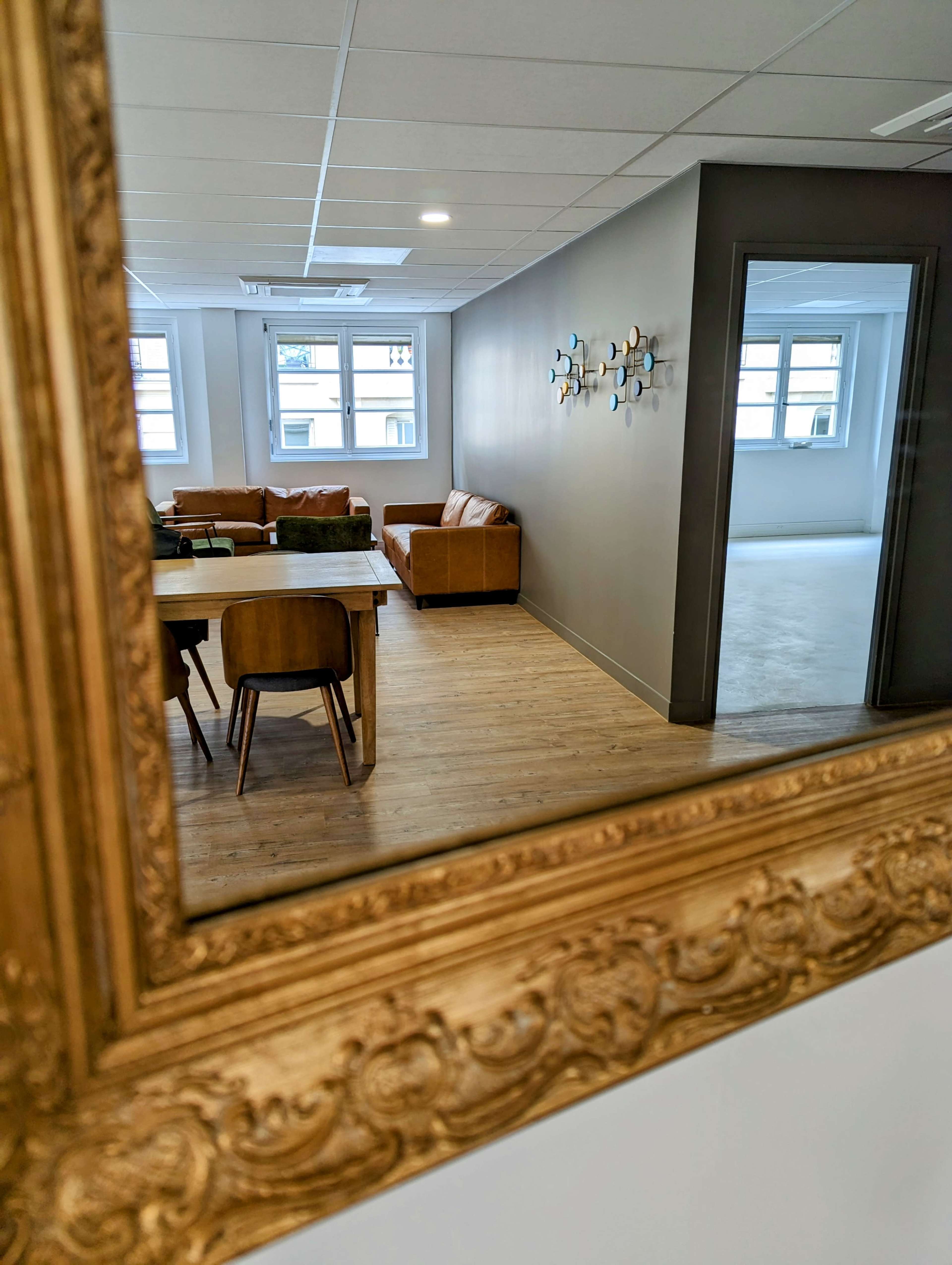 A elegantly framed mirror reflects a cozy lounge area with a brown sofa, wooden table, and chairs in a well-lit, minimalist room.