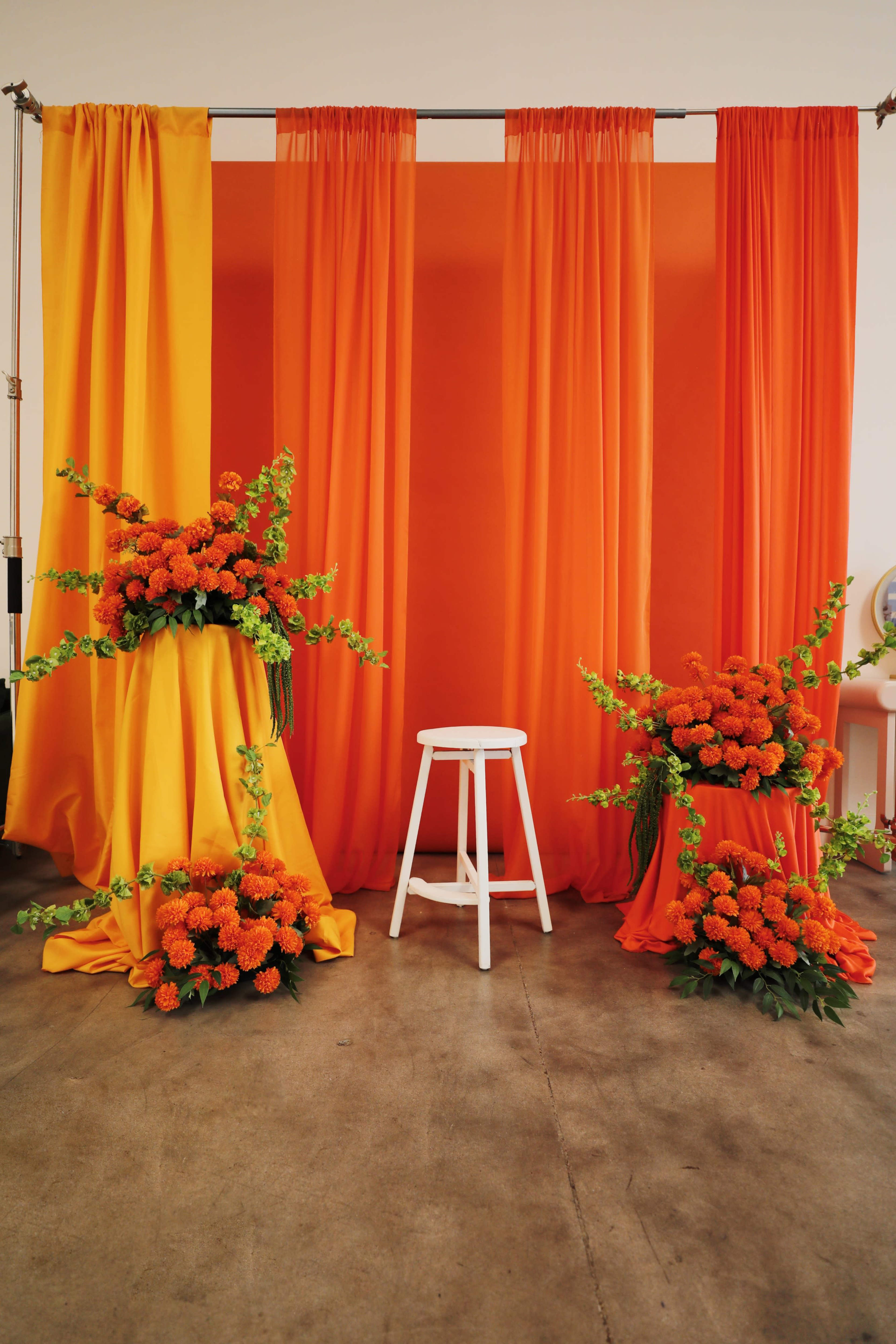 The image features two arrangements of bright orange flowers on draped yellow and orange fabric backgrounds, with a white stool positioned between them.