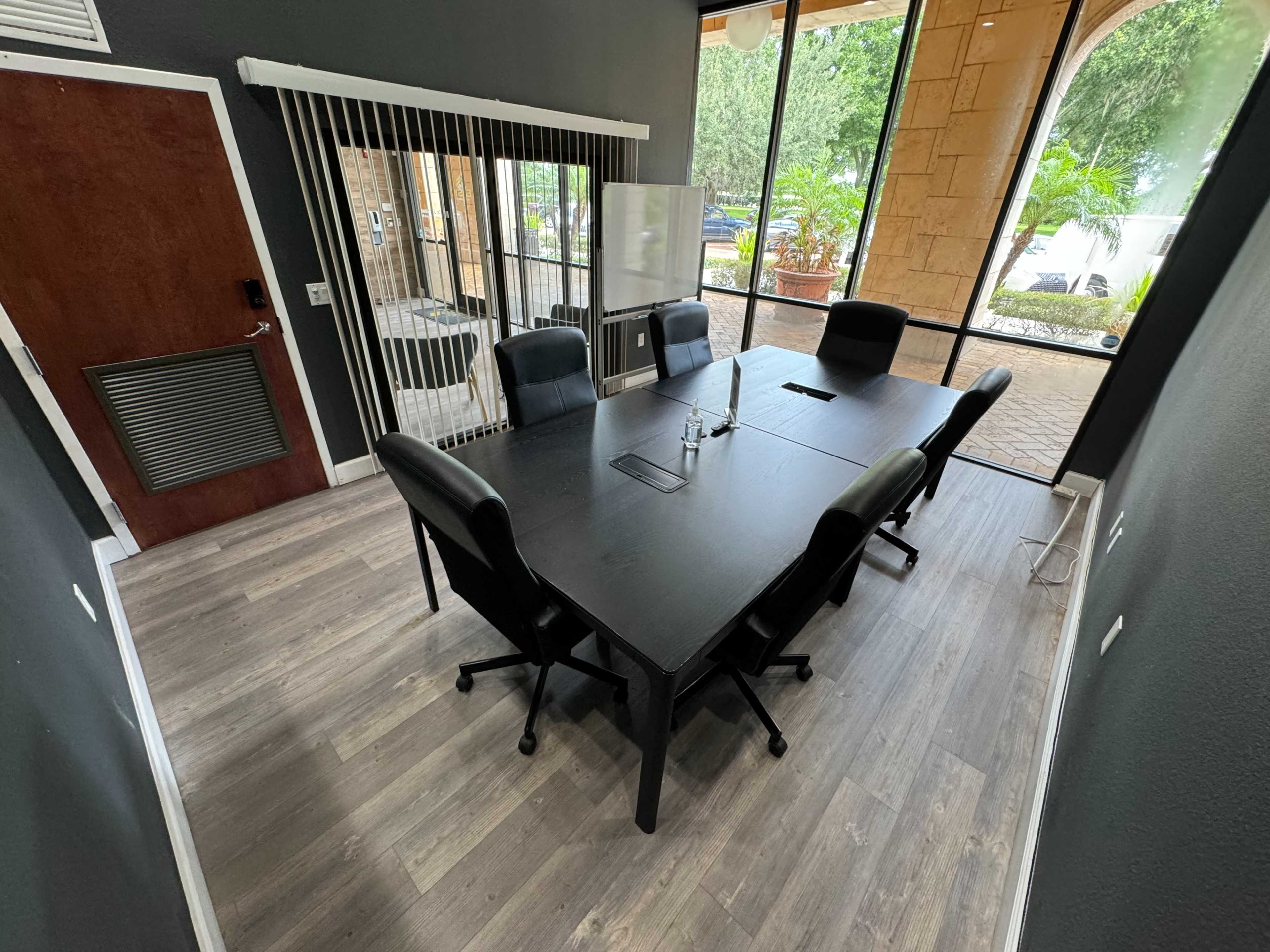 A conference room with a large black table surrounded by six chairs, featuring large windows that provide natural light and a view of greenery outside.