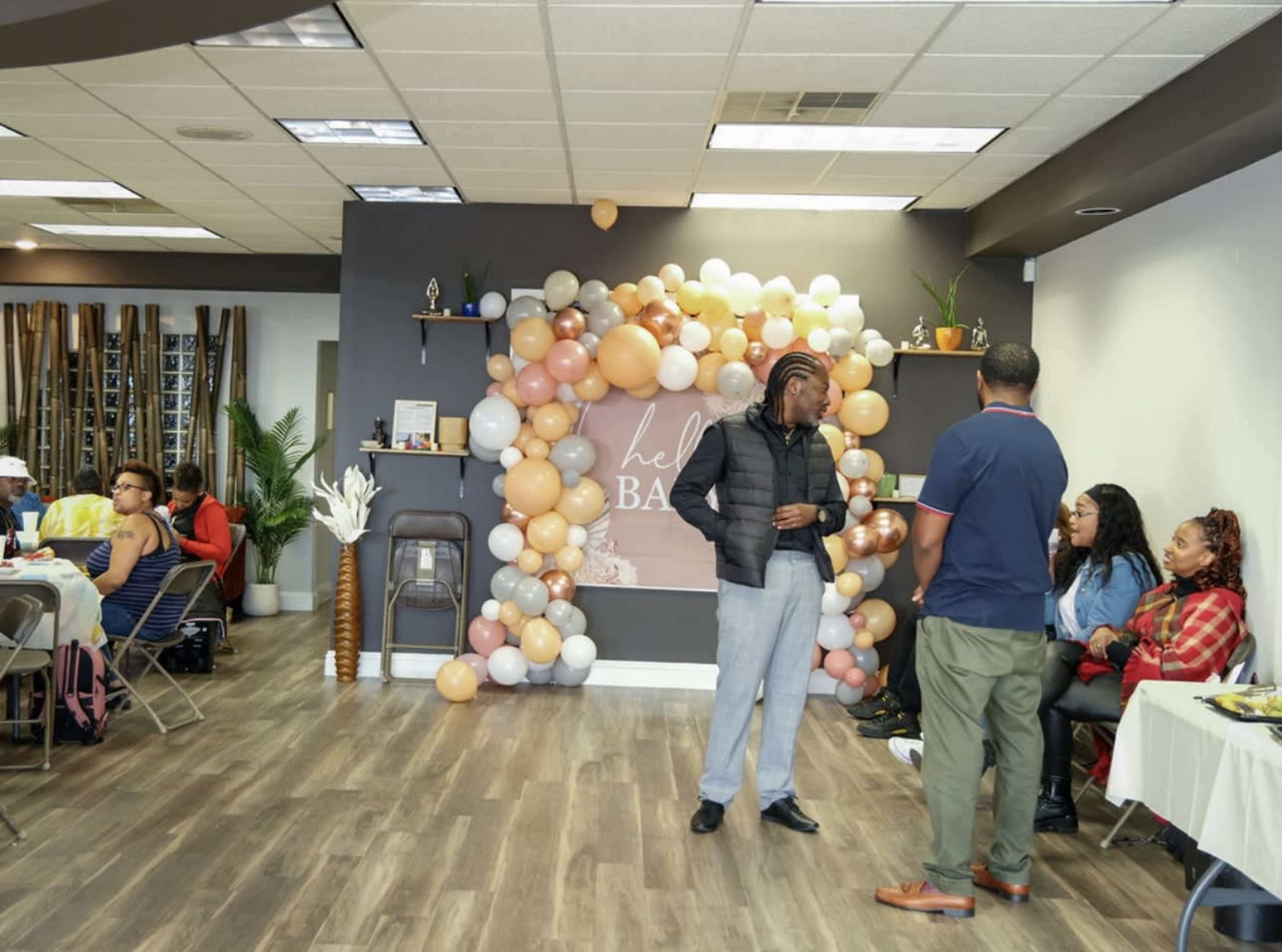 The image shows a lively indoor event space decorated with a balloon backdrop, where people are seated and engaged in conversation.
