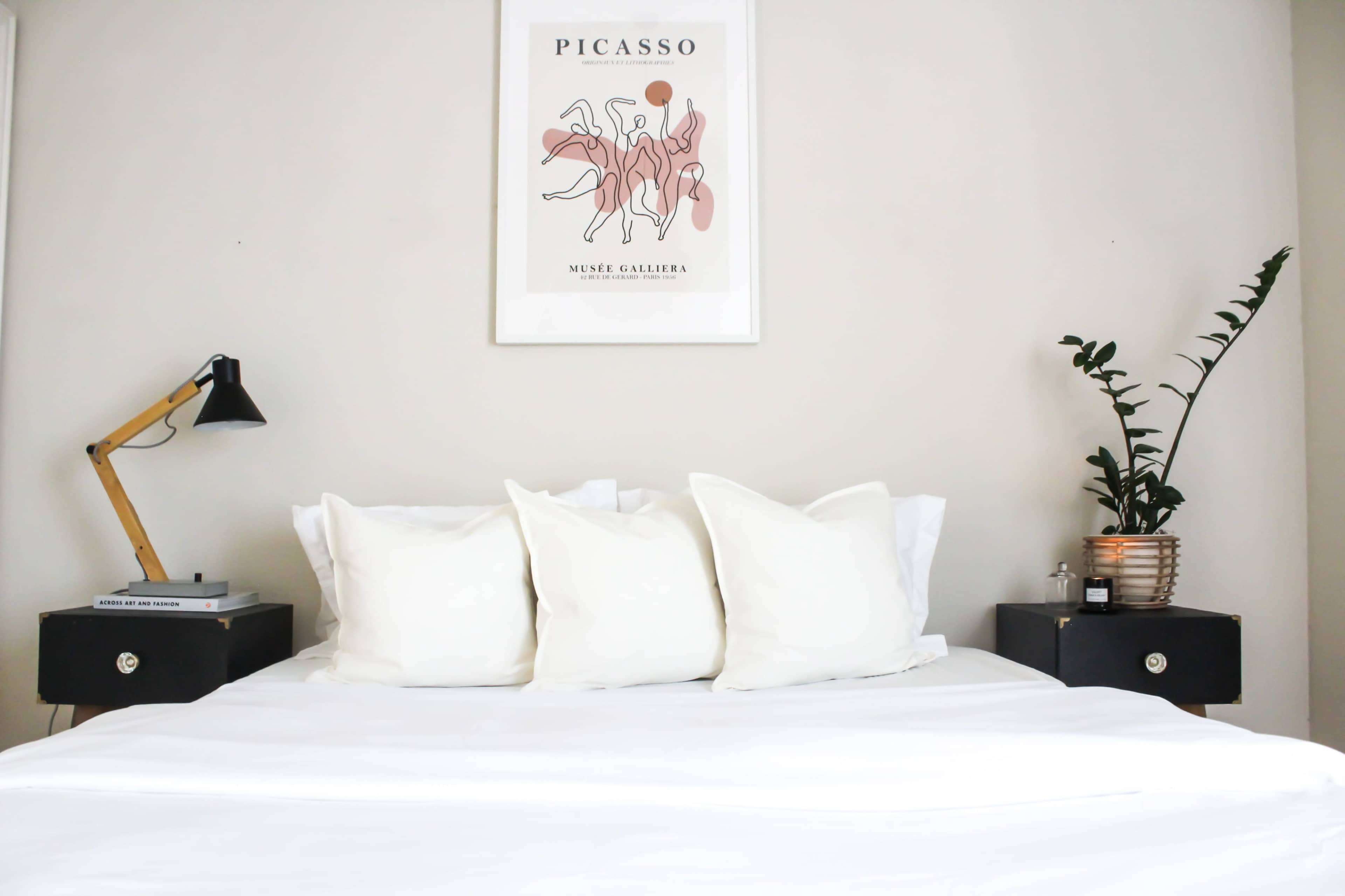 A neatly made bed with white linens and four decorative pillows is flanked by two black nightstands, one featuring a lamp and the other a potted plant.