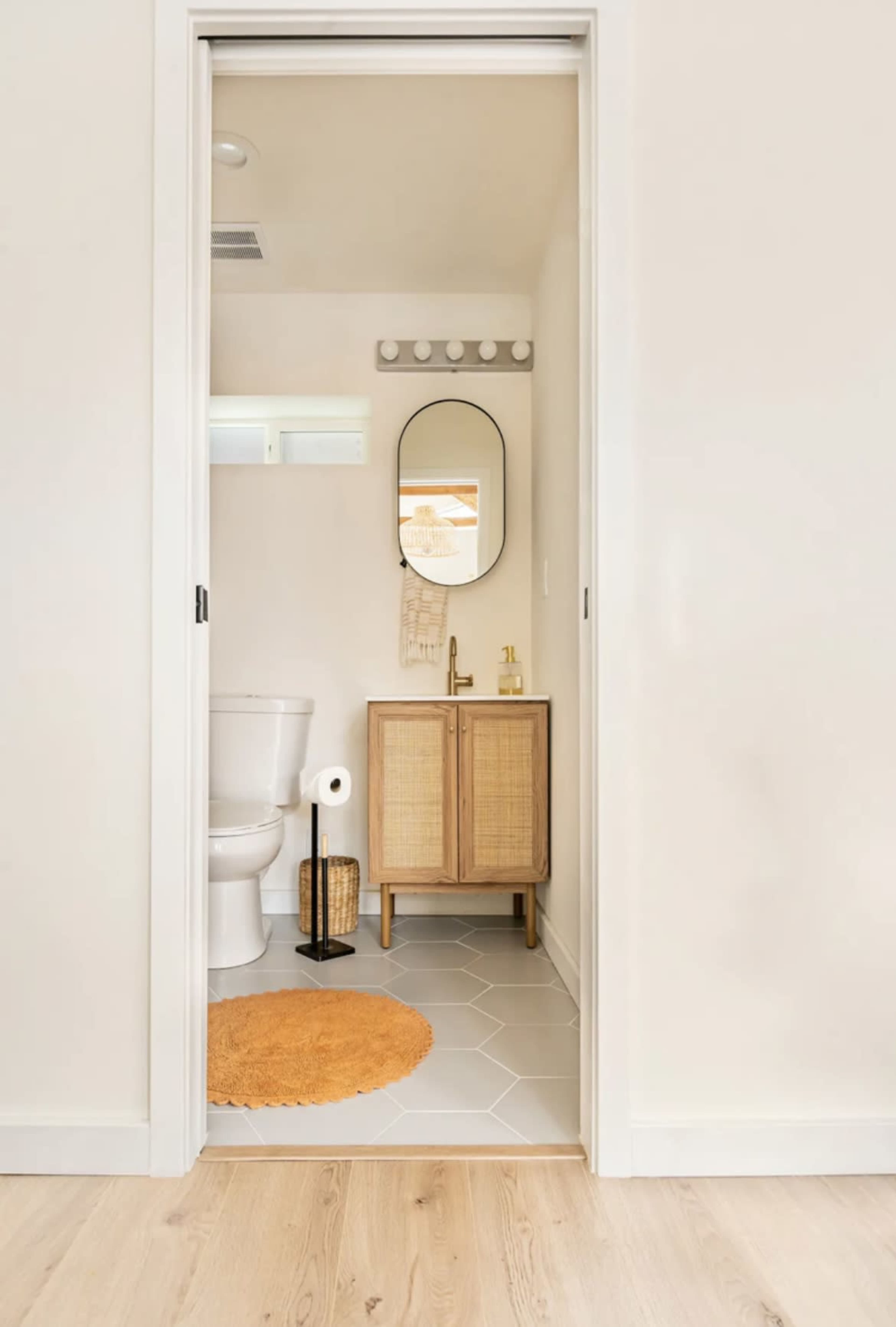 The image shows a bathroom doorway with a wooden cabinet, a round mirror, and a toilet, alongside a brown circular rug on the floor.