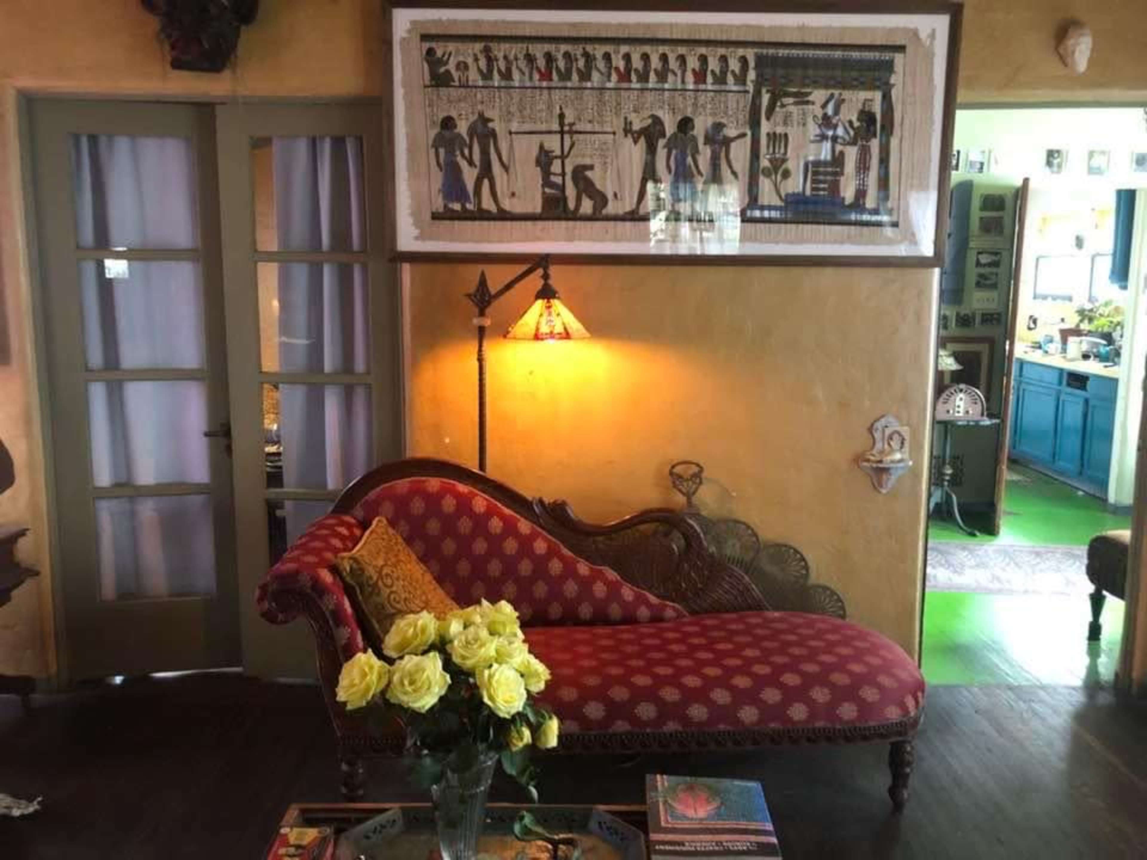 A living room with a red patterned chaise lounge, a floral arrangement of yellow roses on a coffee table, and a wall adorned with an Egyptian-themed artwork.
