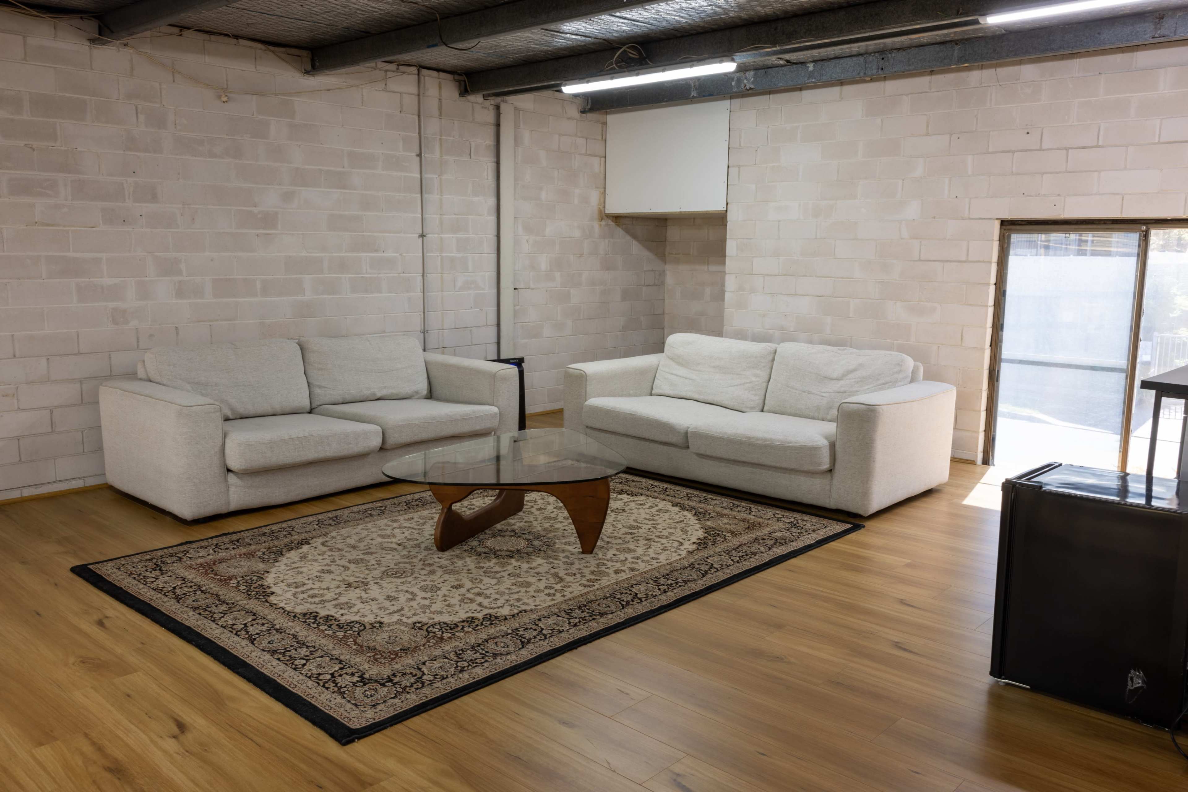 A spacious room features two light-colored sofas facing a glass coffee table on a patterned rug, with a large window allowing natural light.
