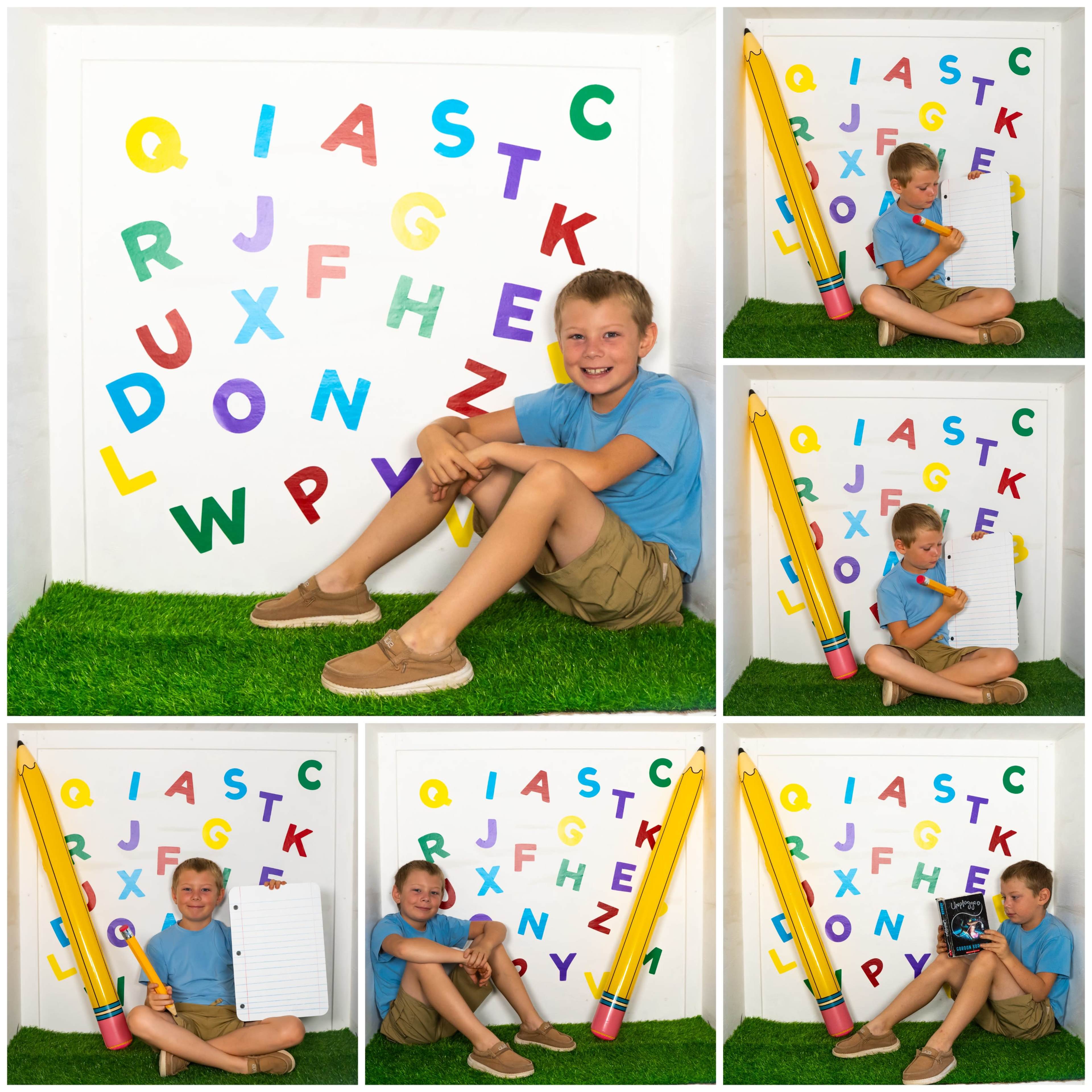A boy in a blue shirt and khaki shorts sits on grass in front of a colorful alphabet display, holding a large notepad and leaning against a giant pencil.