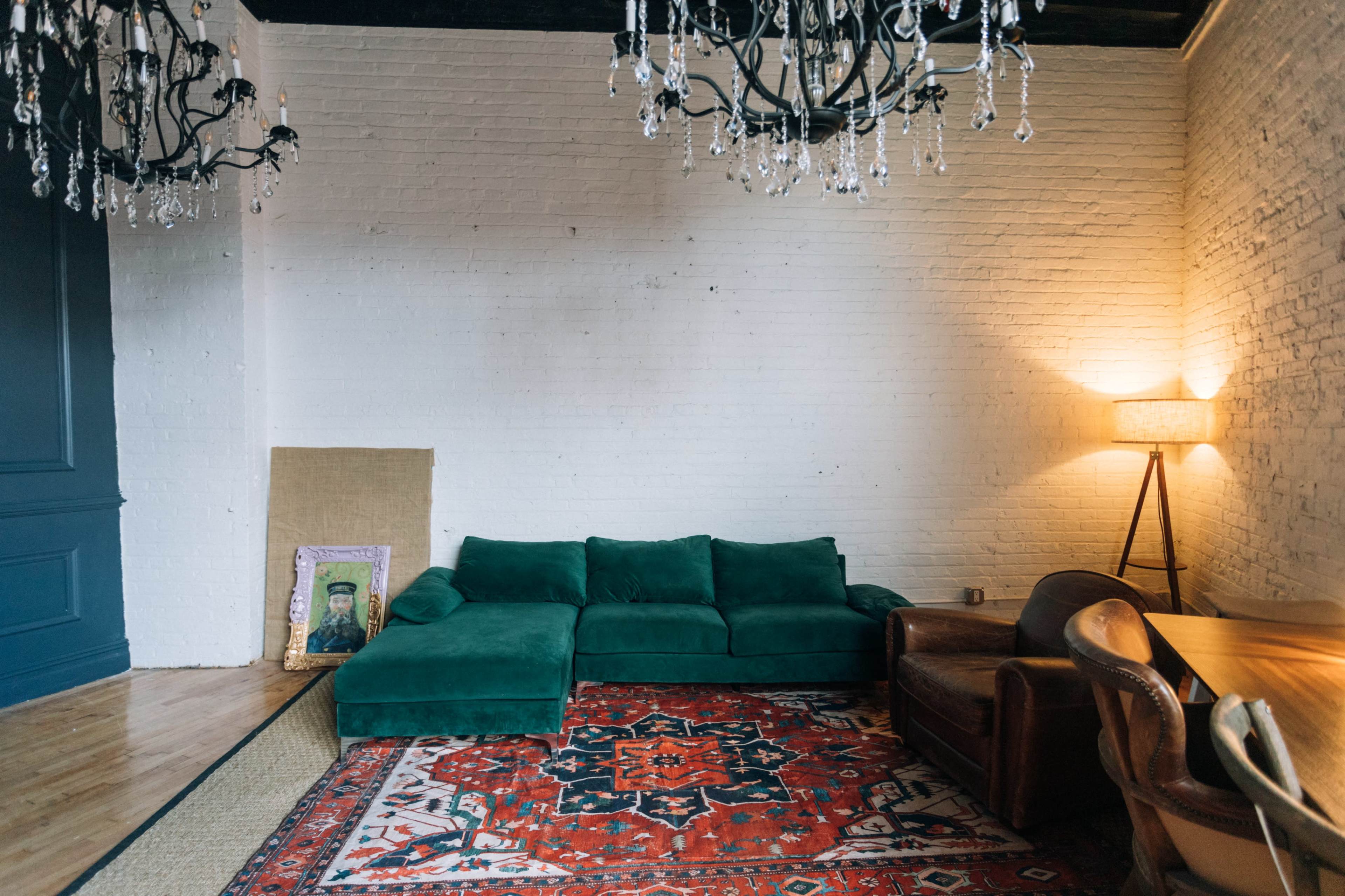 The image shows a minimalist living room with a green sectional sofa, a decorative rug, a lamp, and two chandeliers hanging from the ceiling.