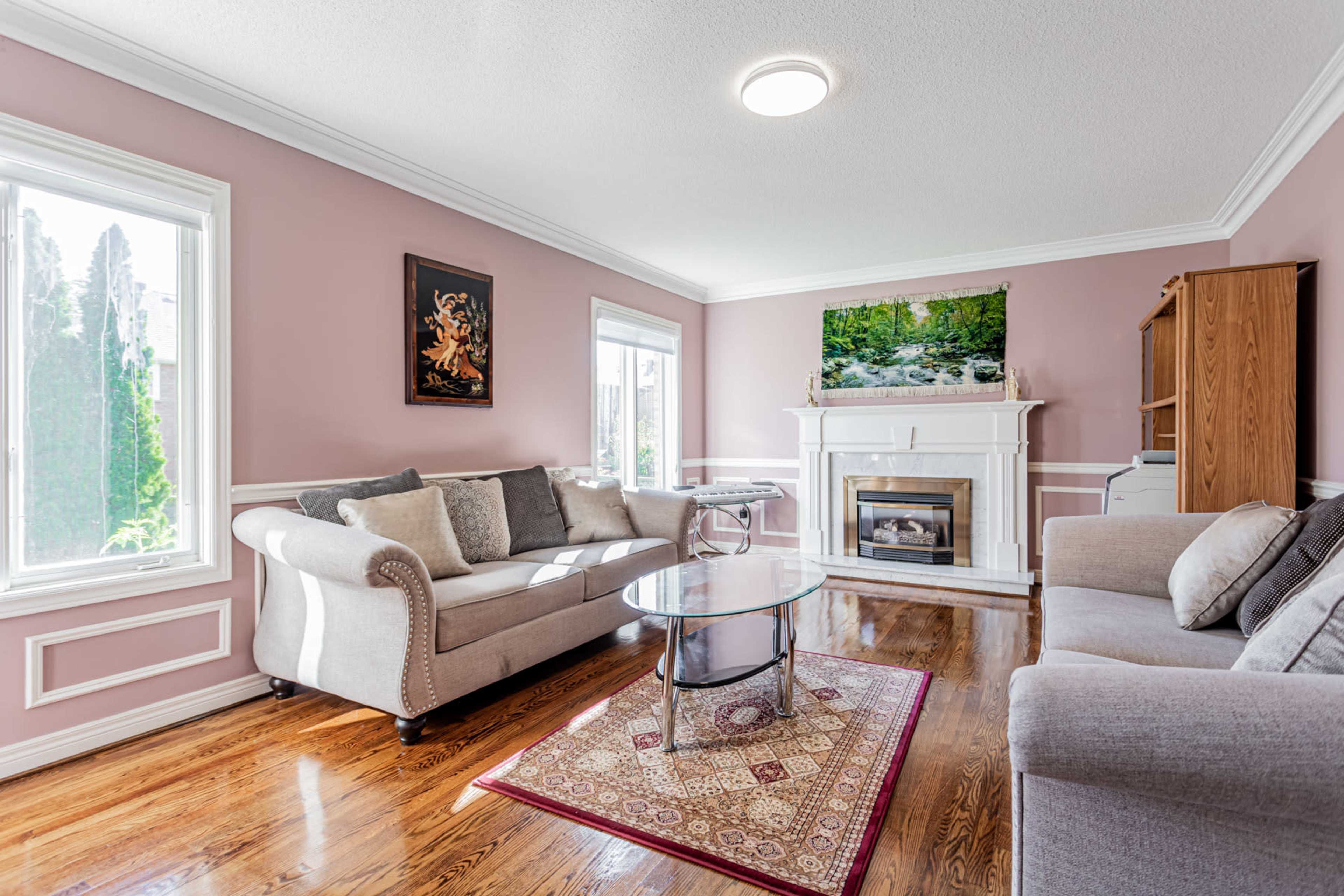 A well-furnished living room with a fireplace and large windows, featuring a light purple wall color and wooden flooring.