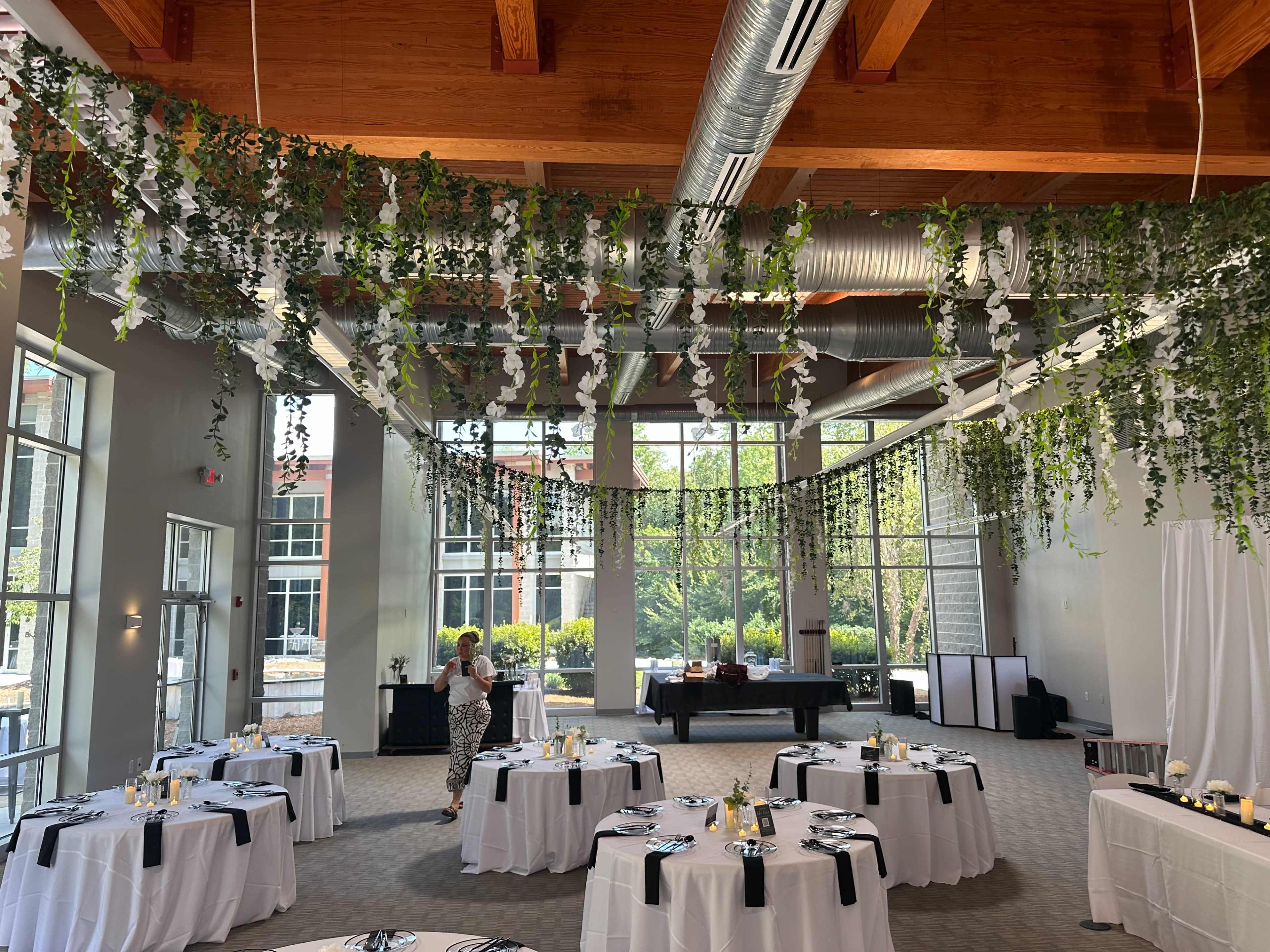 The image shows a decorated indoor venue with round tables, each set with white tablecloths and black accents, beneath an overhead arrangement of greenery and flowers.