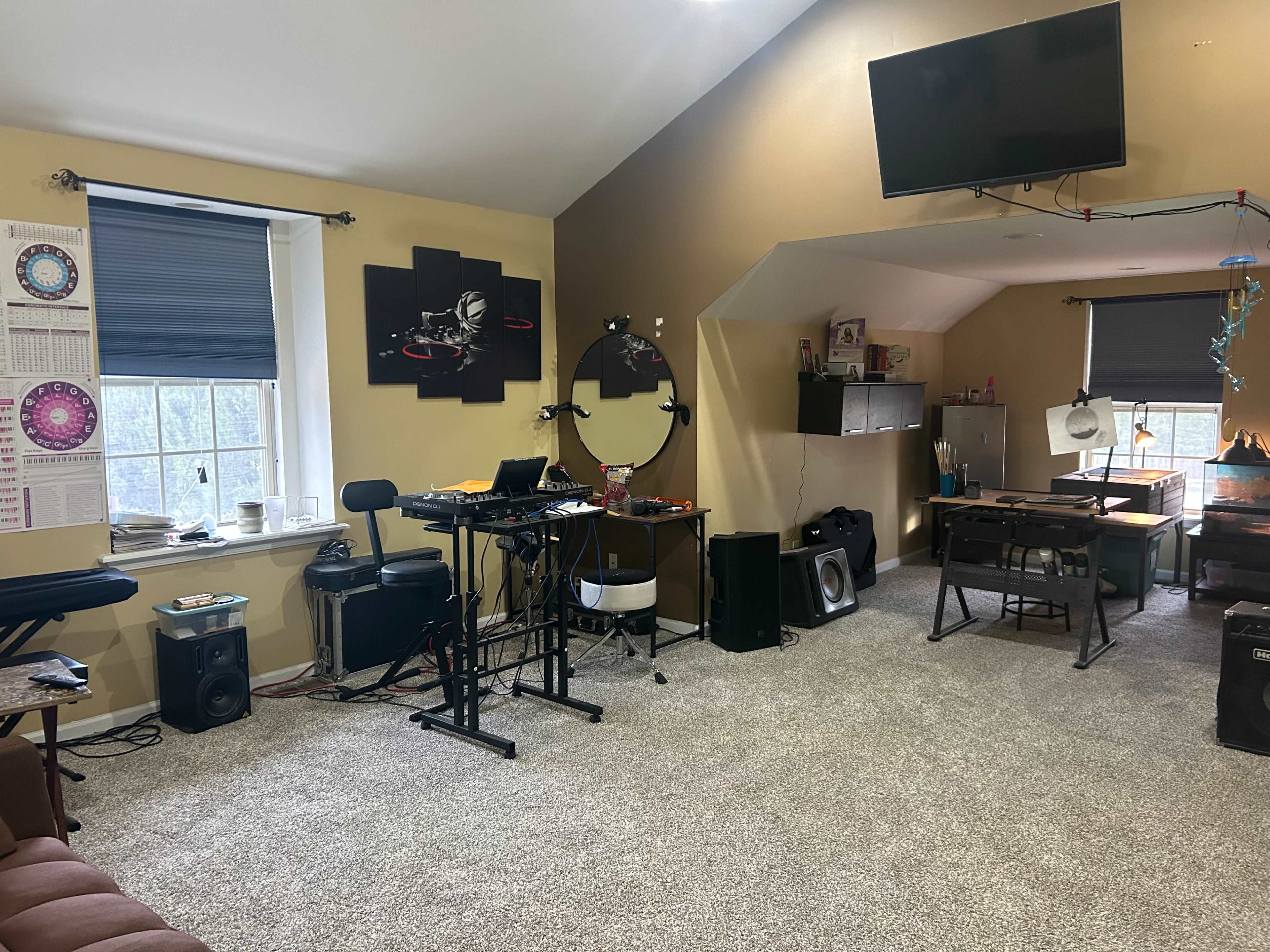 The image shows a spacious room with a workspace that includes a keyboard, electronic equipment, and a seating area, all set against a backdrop of light-colored walls and carpet.