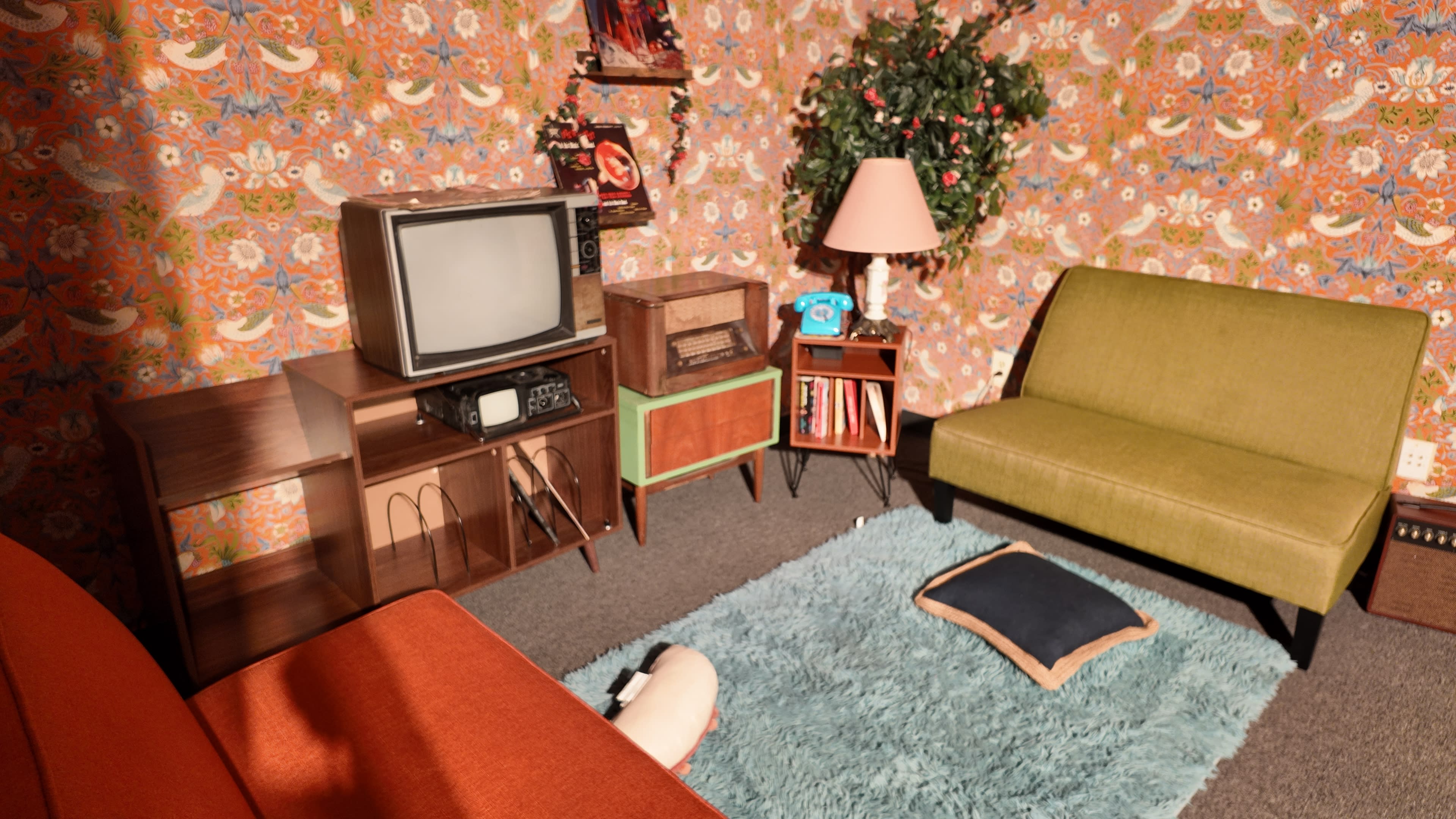 The image shows a vintage living room with green and orange furniture, a classic television, and a decorative floral wallpaper.