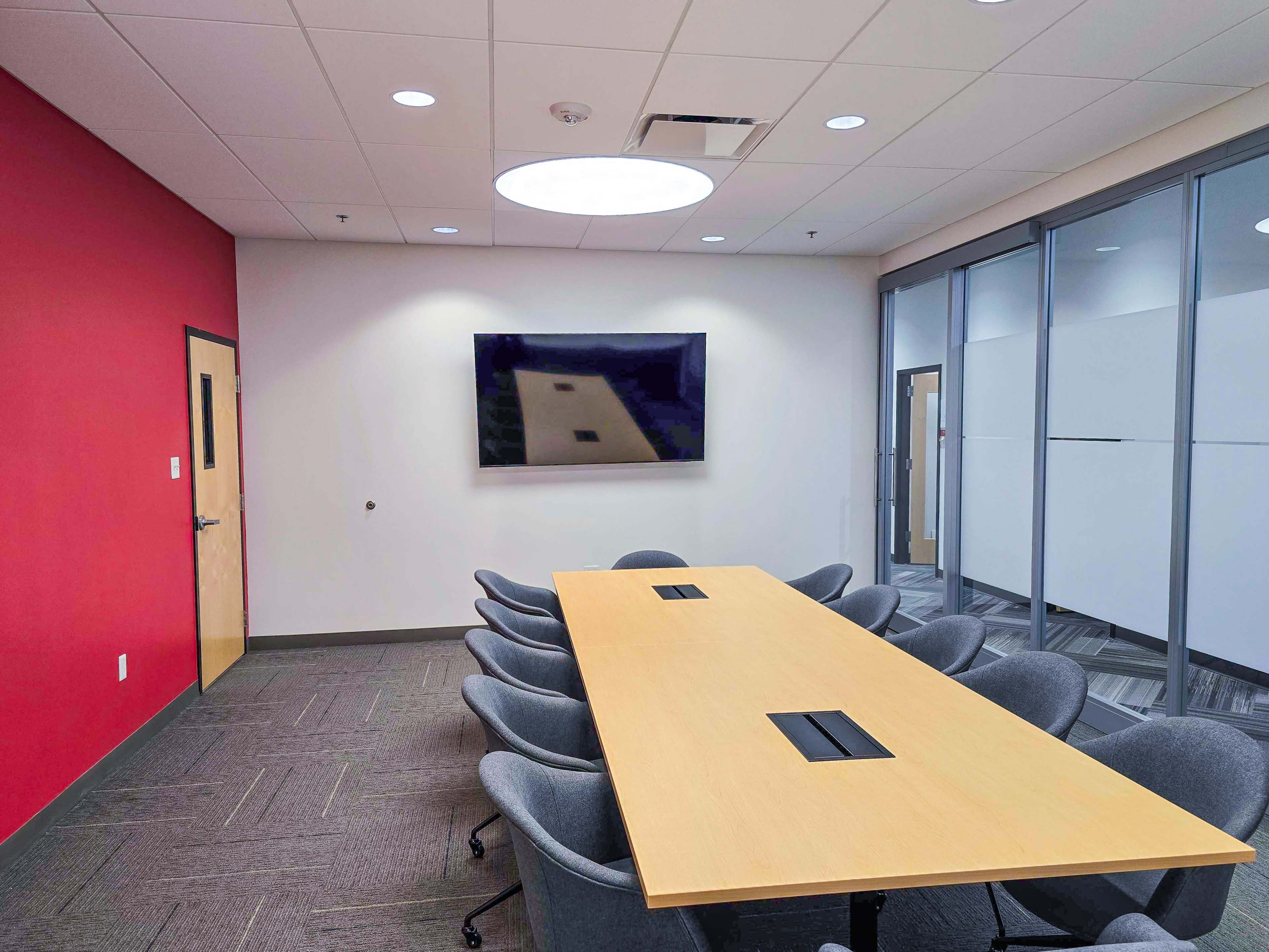 The image shows a modern conference room featuring a long wooden table surrounded by gray chairs, with a large screen mounted on a wall and a circular light fixture above.
