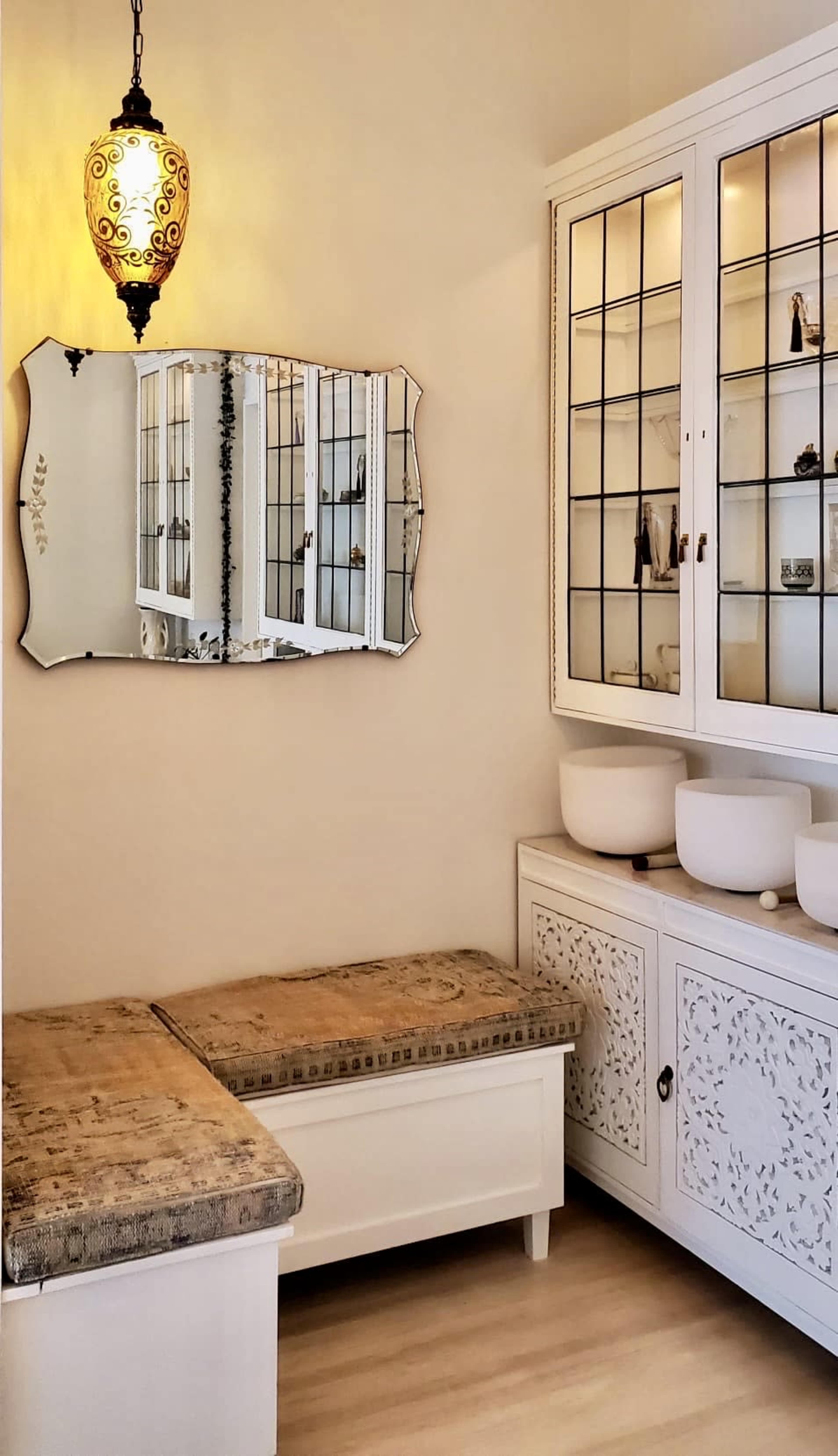The image shows a serene room featuring a mirrored wall ornament, a decorative light fixture, and a white cabinet with glass doors displaying items inside.