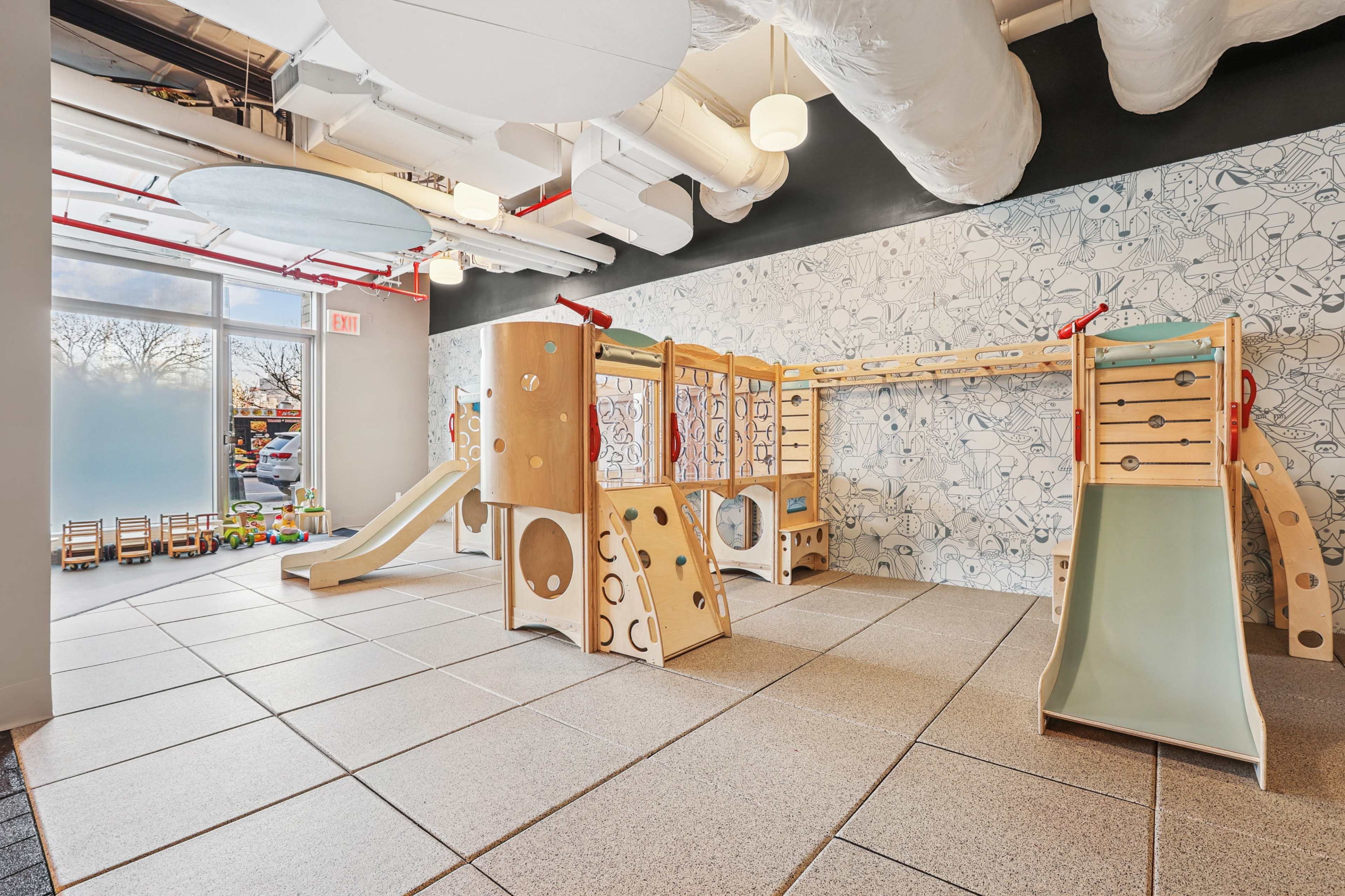 The image shows an indoor play area featuring wooden climbing structures, slides, and a textured floor in a brightly lit space.