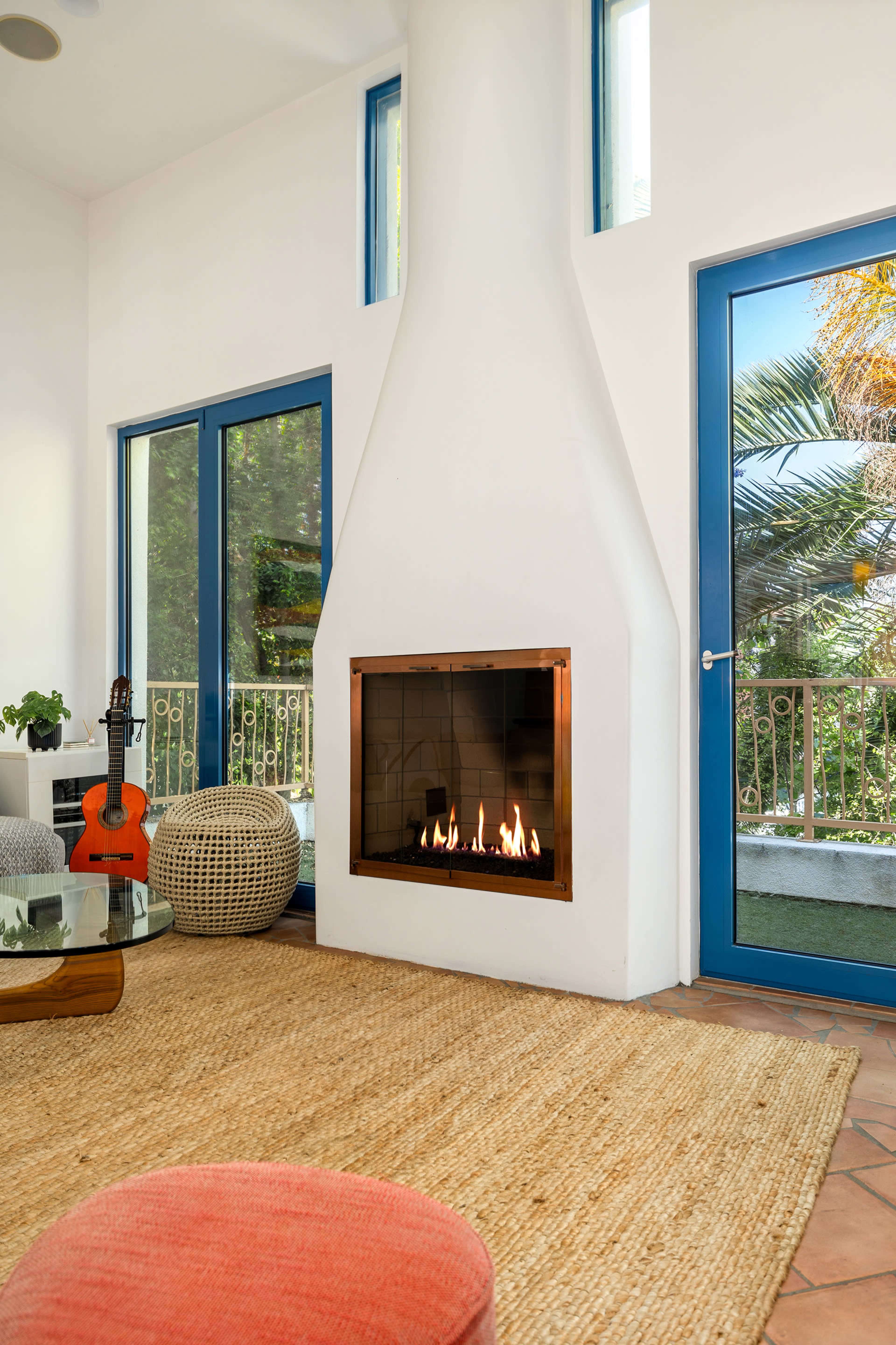 A modern living room features a central fireplace with a glass front, surrounded by large blue windows and a circular jute rug on the terracotta floor.