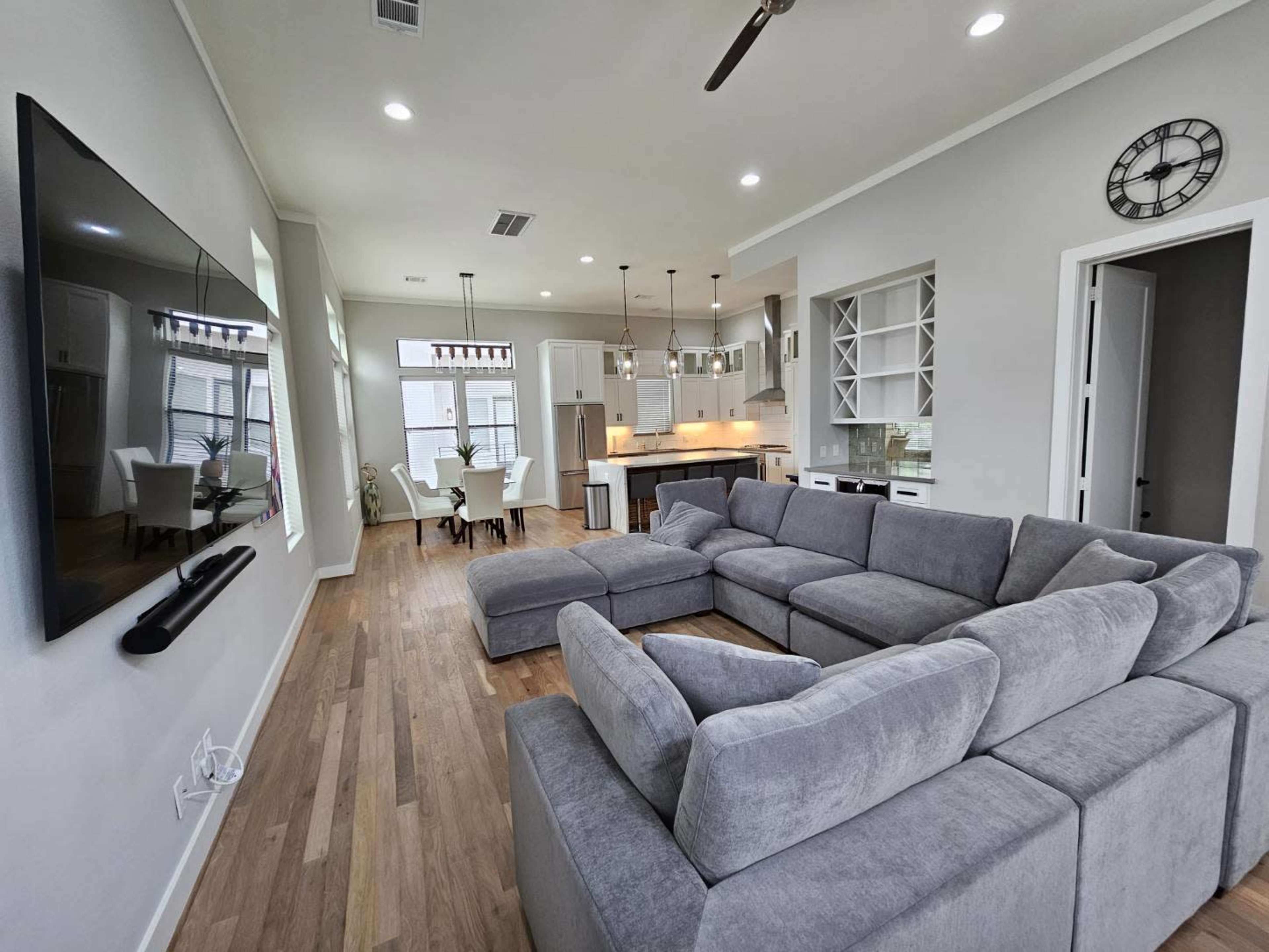 A modern living room with a large sectional sofa facing a wall-mounted television, leading into an open kitchen area with a dining table.