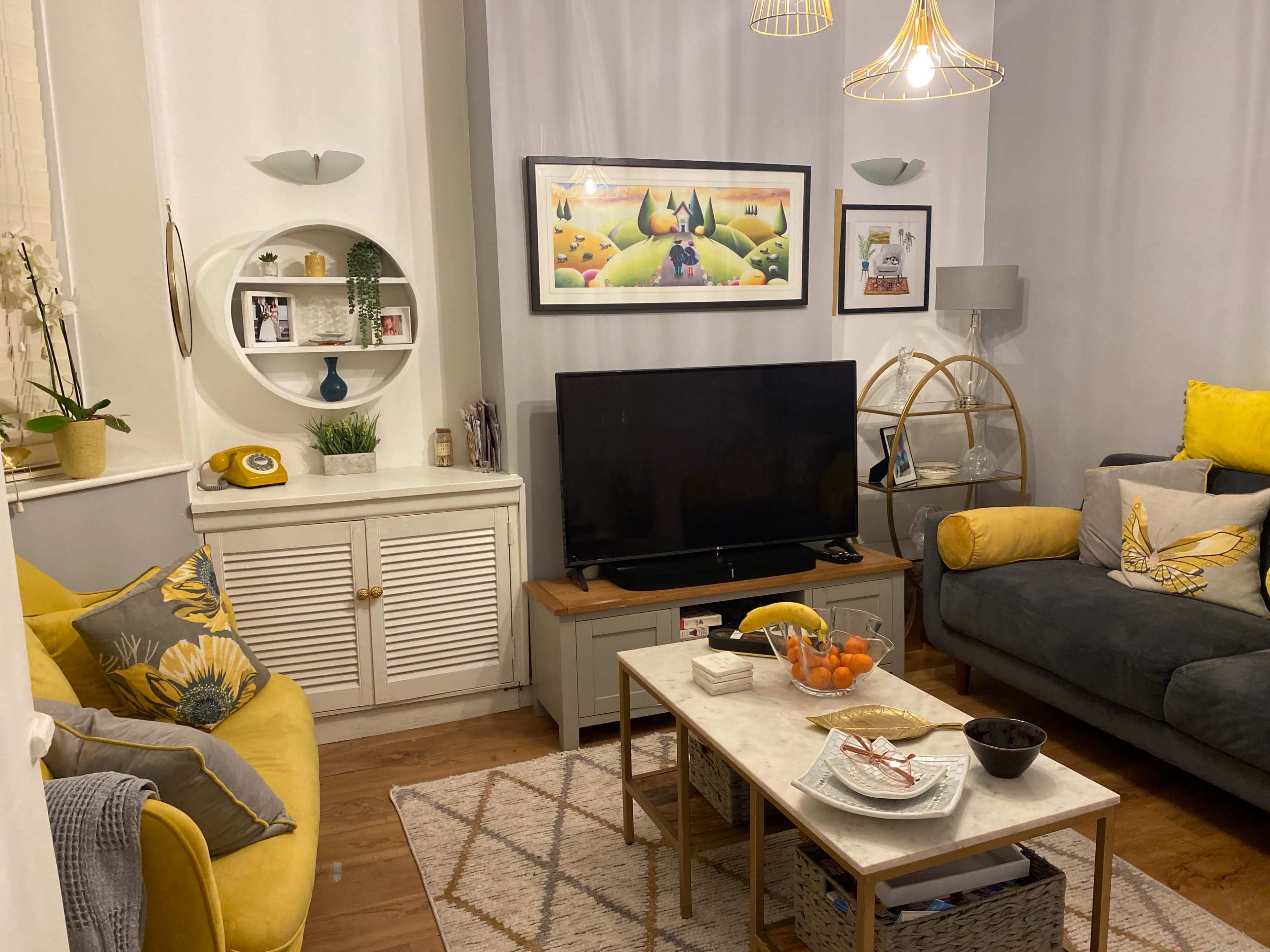 A cozy living room with a gray sofa, a yellow chair, a television on a wooden stand, and a coffee table with snacks, surrounded by decorative elements like plants and artwork.