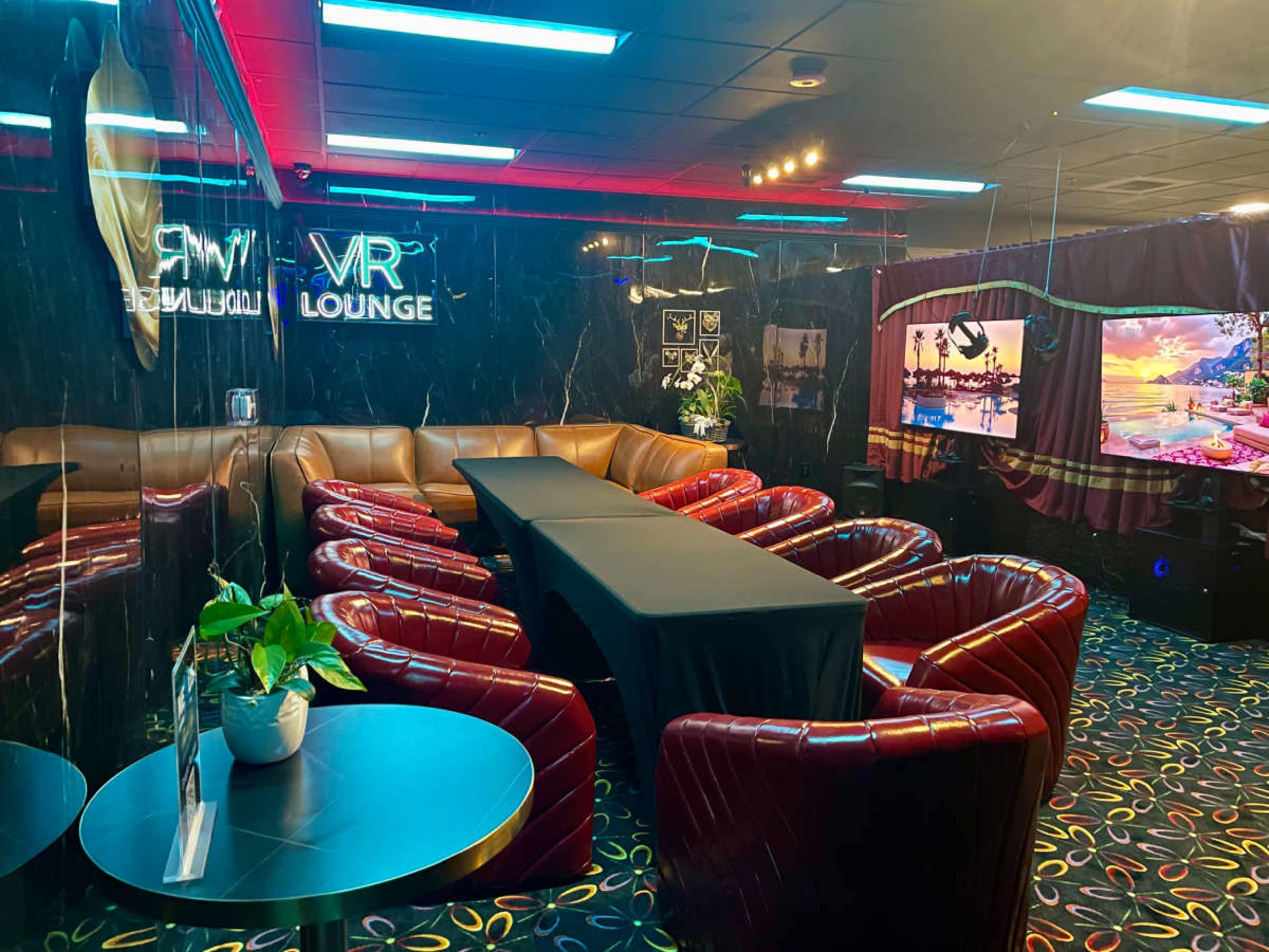 The image showcases a modern lounge designed for virtual reality experiences, featuring red leather chairs, a long table, and large screens displaying vibrant images.