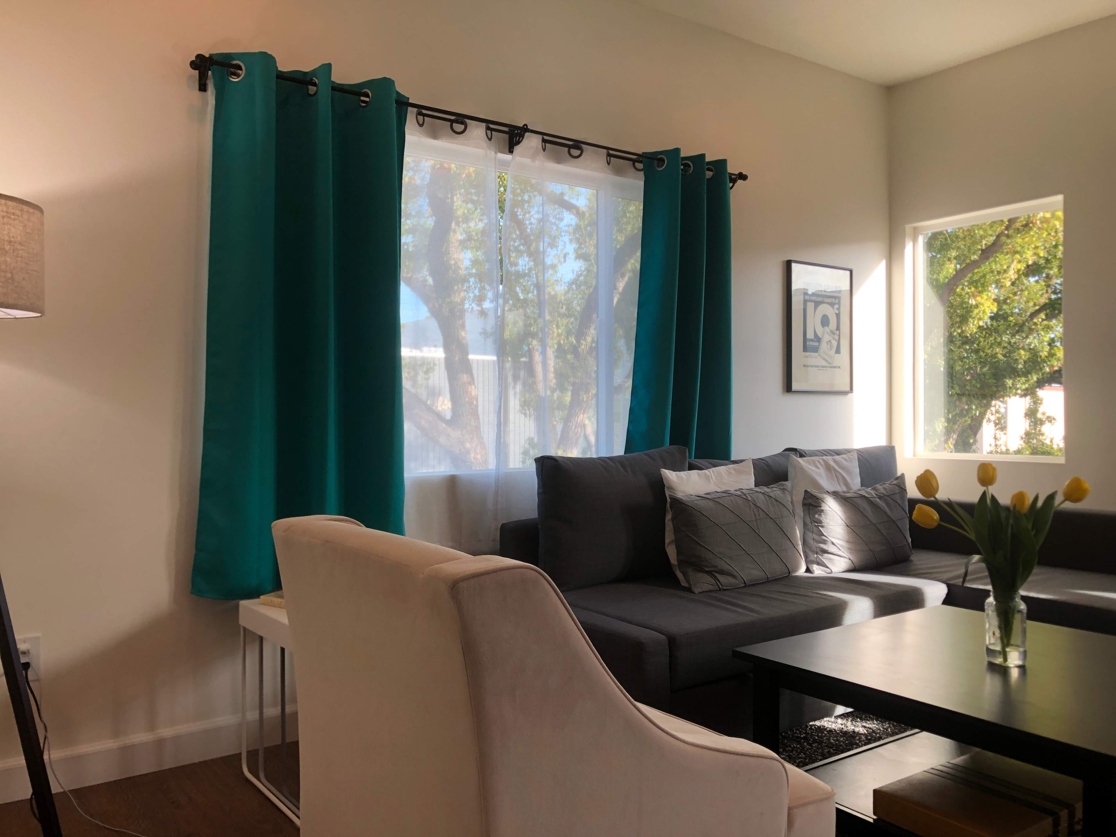 The image shows a bright living room with a gray sofa, large windows draped with turquoise curtains, and a small table with a vase of yellow flowers.