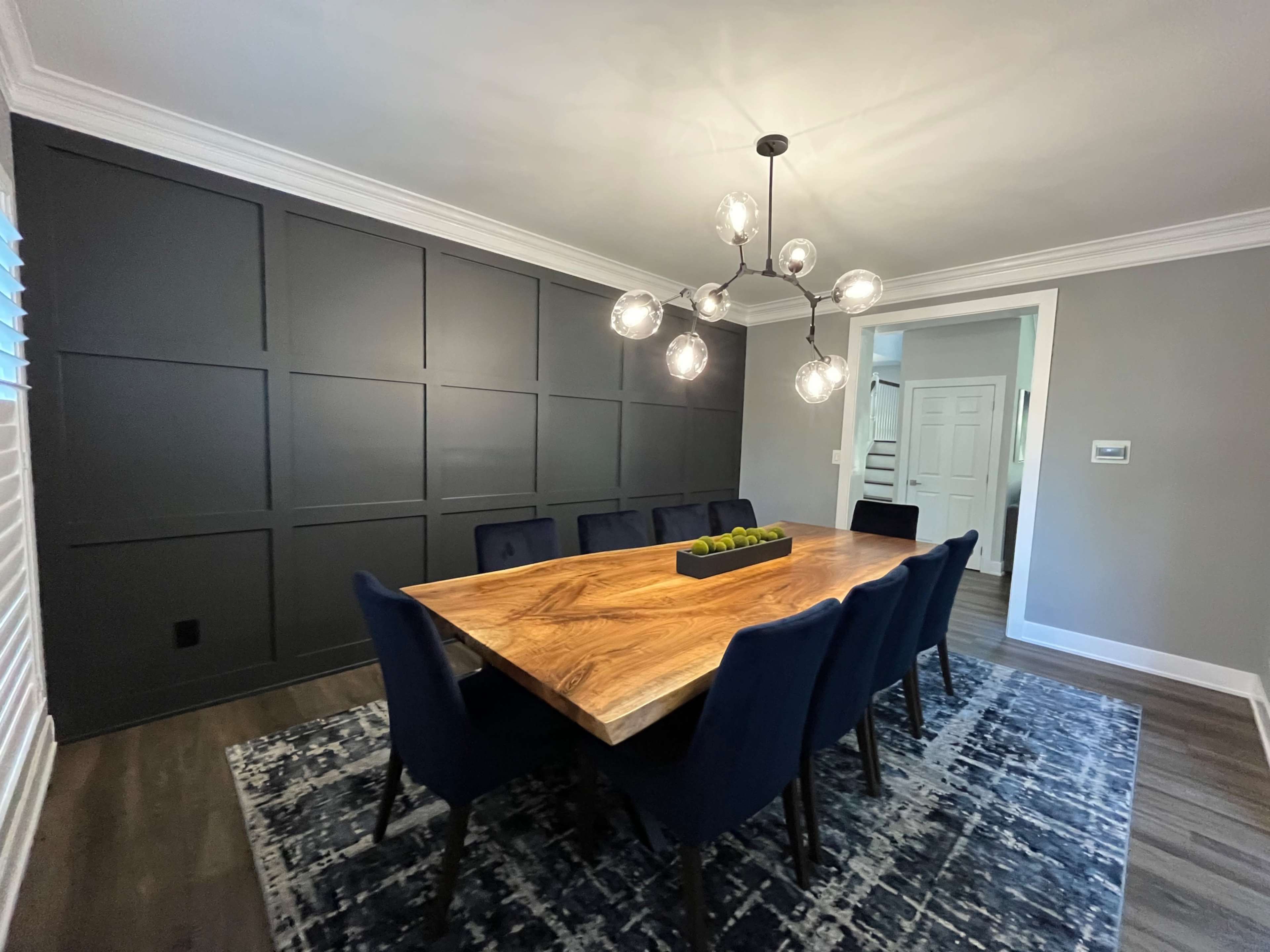 A spacious dining room features a long wooden table surrounded by dark blue chairs, with a modern light fixture overhead and a textured rug beneath.