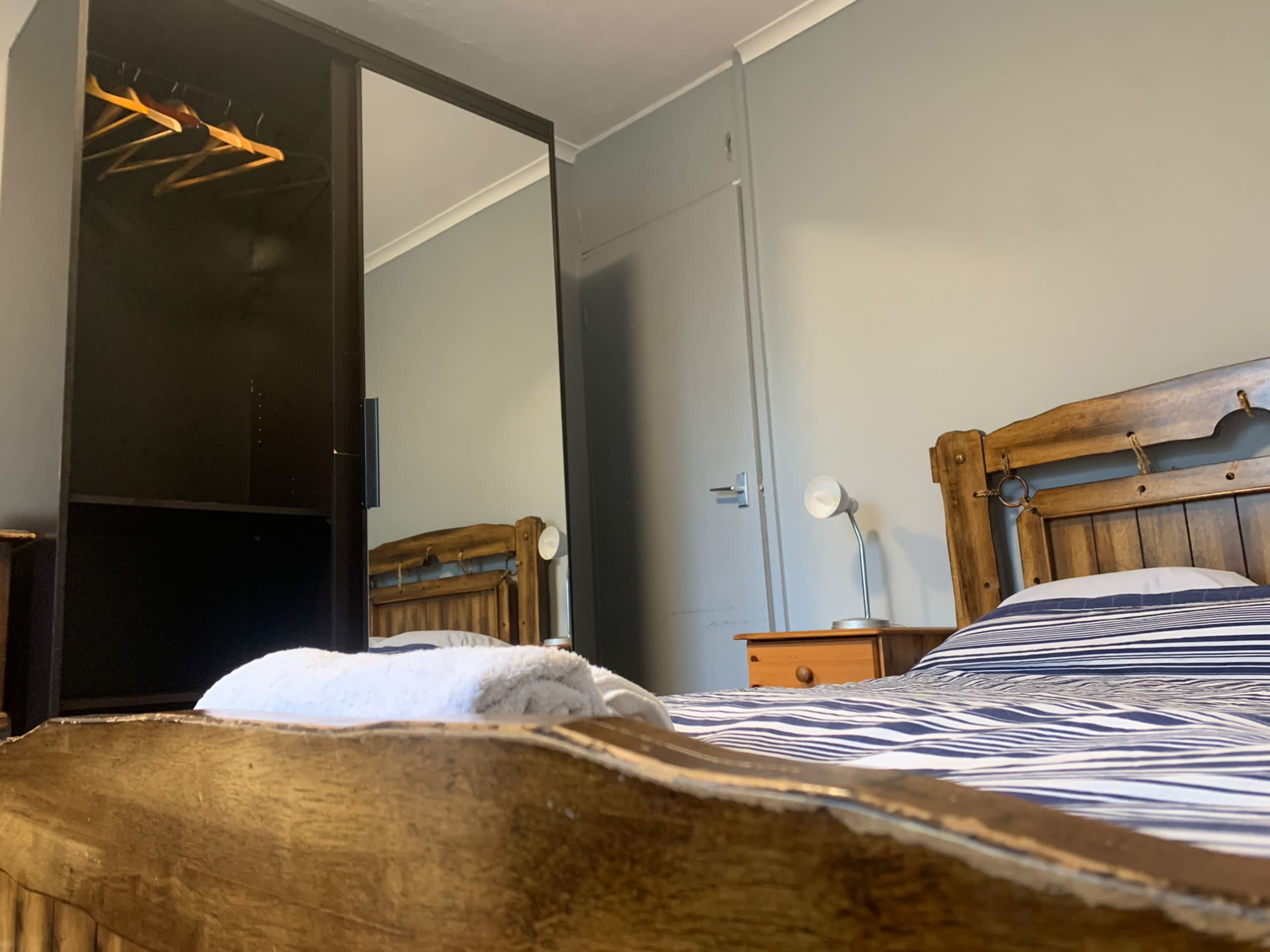 A neatly arranged bedroom features a wooden bed with a striped blanket, a mirror-fronted wardrobe, and a bedside table with a lamp.