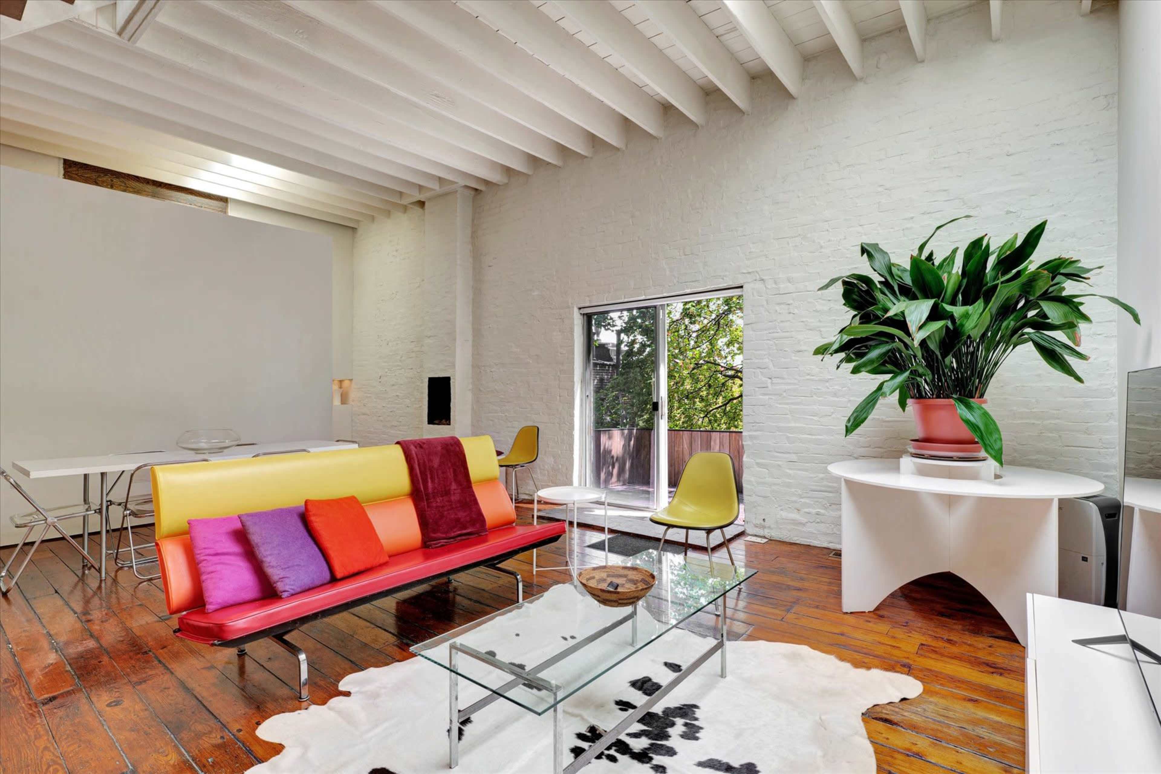 A modern living room features a colorful sofa, a glass coffee table, a potted plant, and large windows that allow natural light to fill the space.