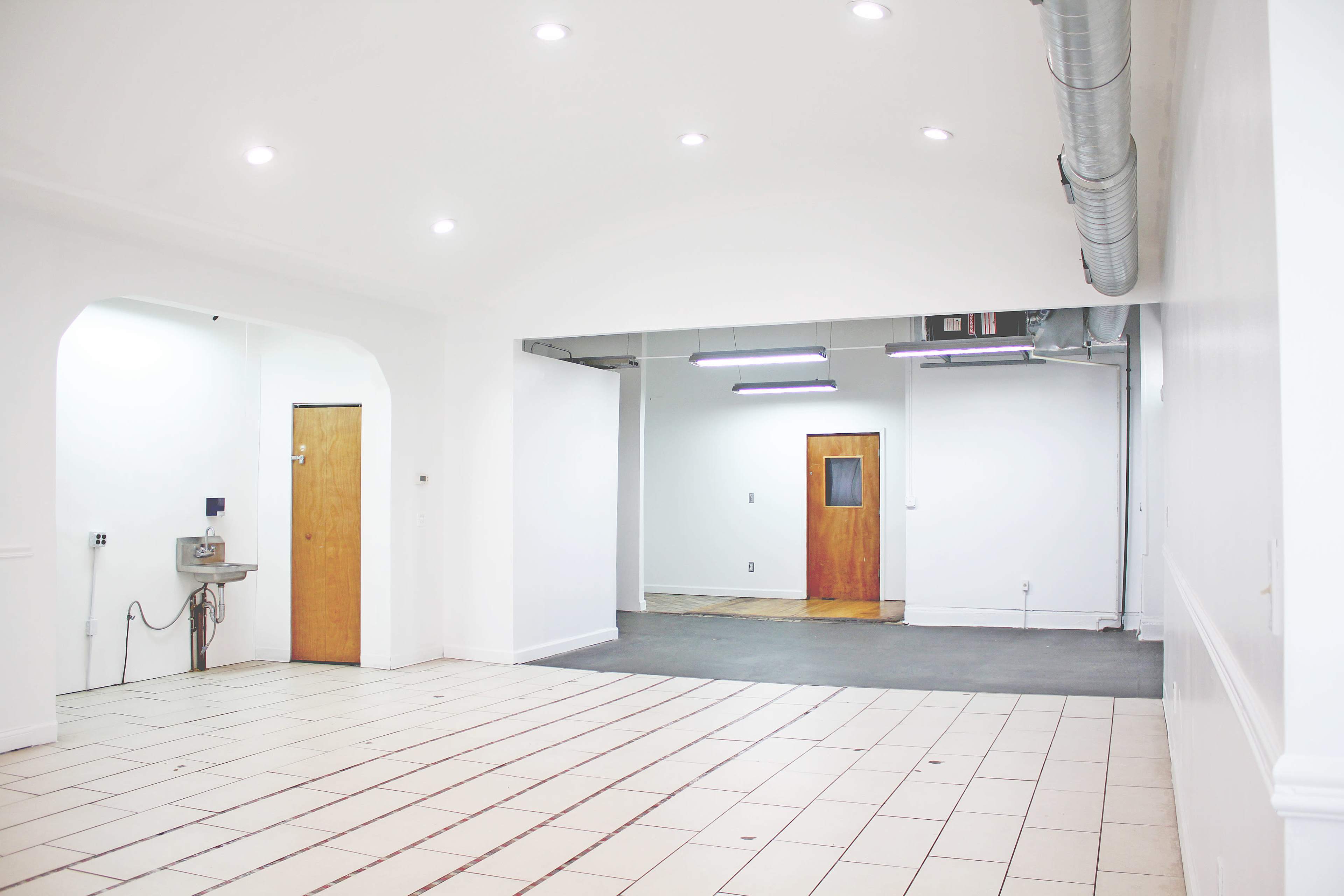 The image shows a spacious, empty room with white walls, tiled flooring, and two wooden doors, one leading to a small area with a sink.