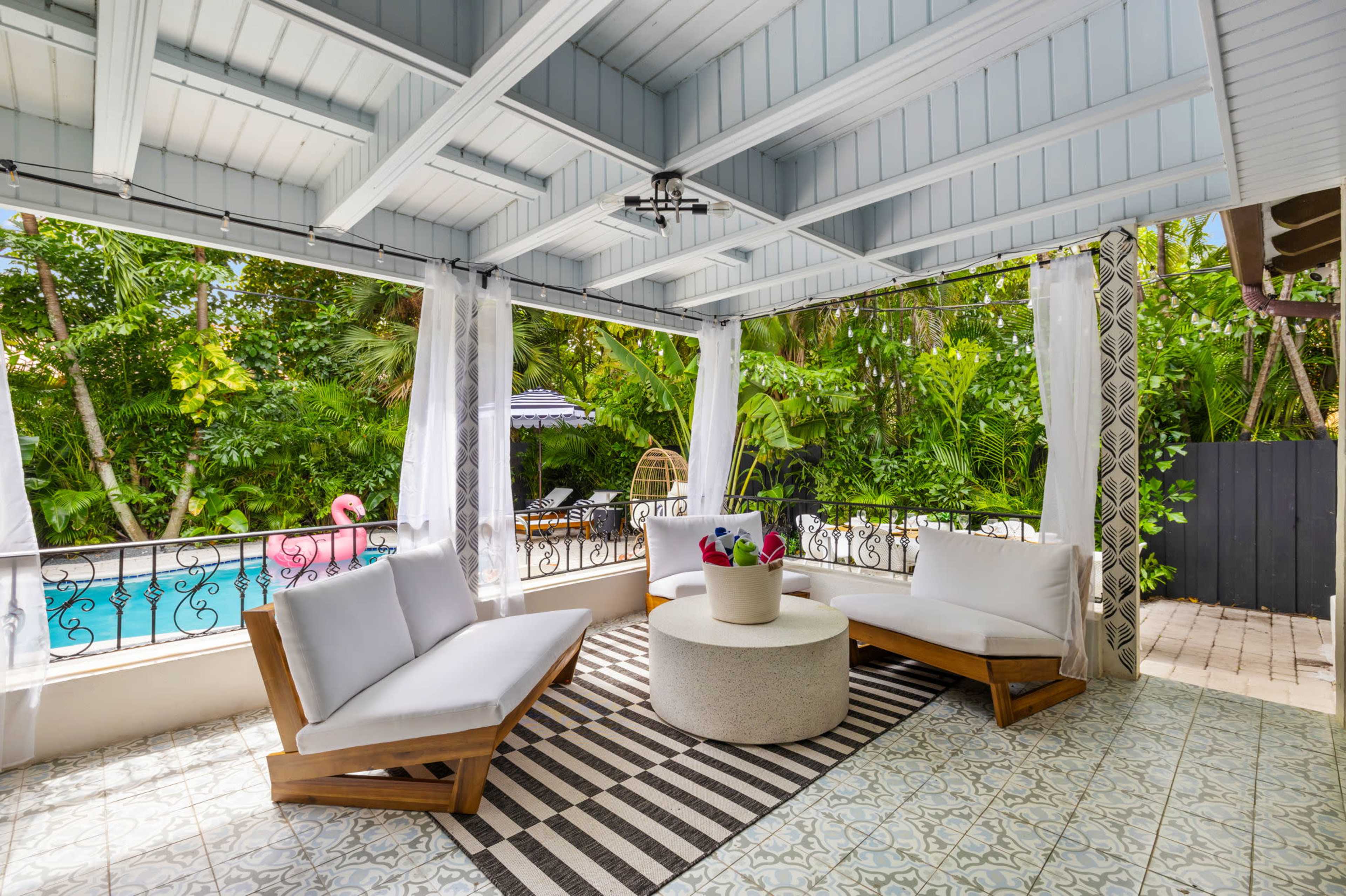 A covered outdoor seating area features two lounge chairs and a circular table, surrounded by lush greenery and a swimming pool visible in the background.