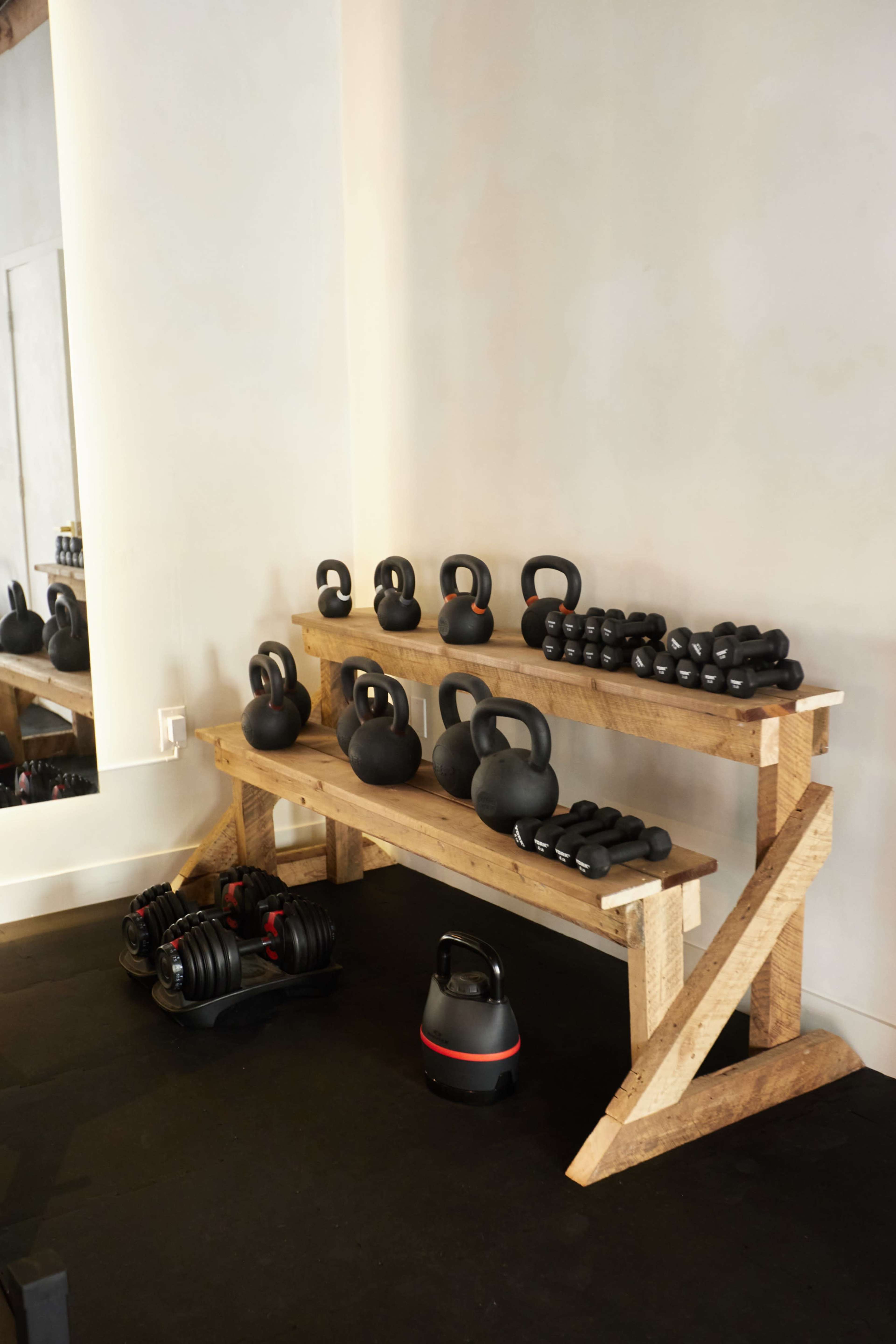 A wooden rack displays an assortment of black kettlebells and dumbbells in a gym setting.