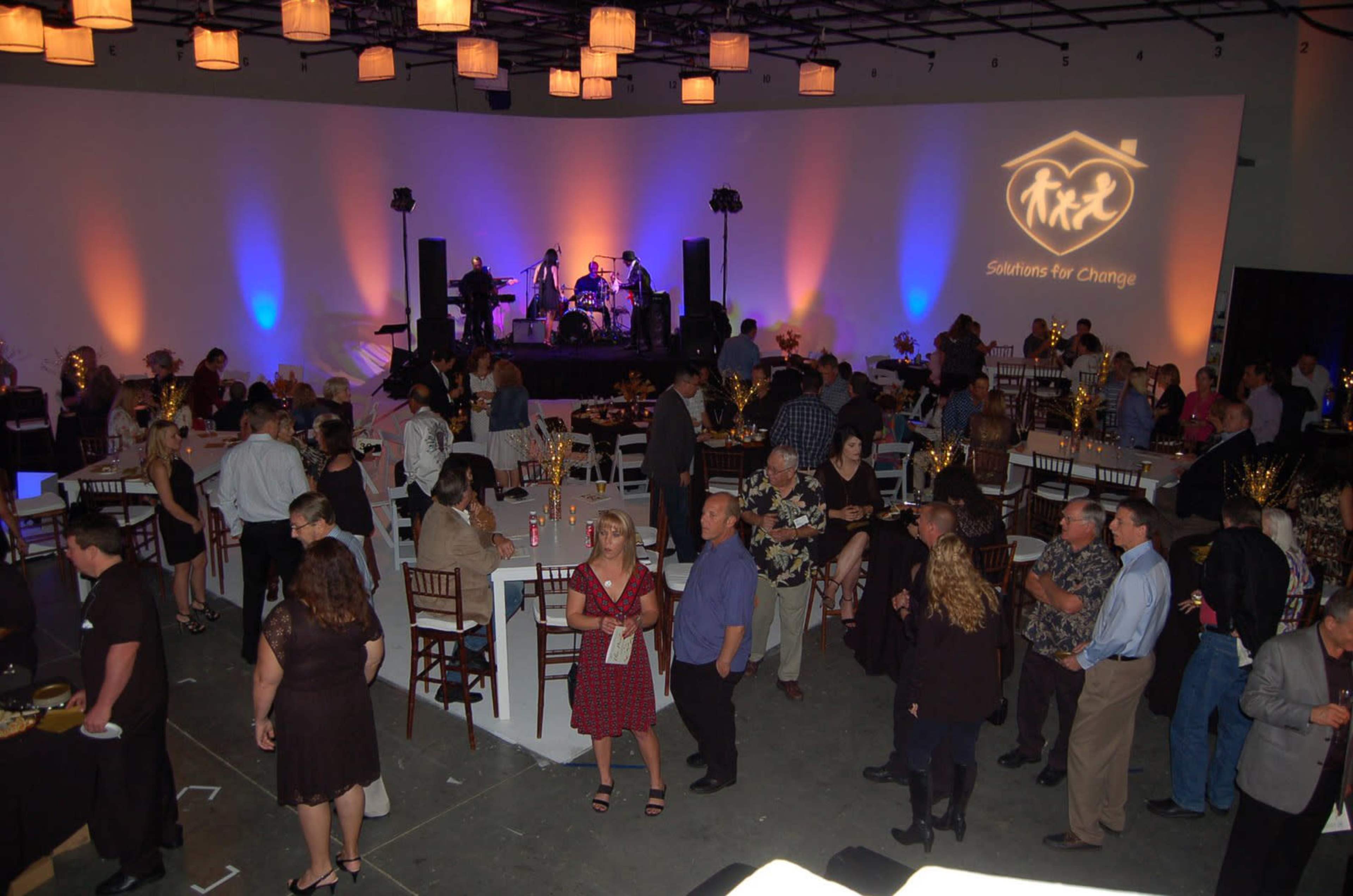 A large crowd gathers in a venue decorated with uplighting, food tables, and a stage where a band performs, while guests socialize.
