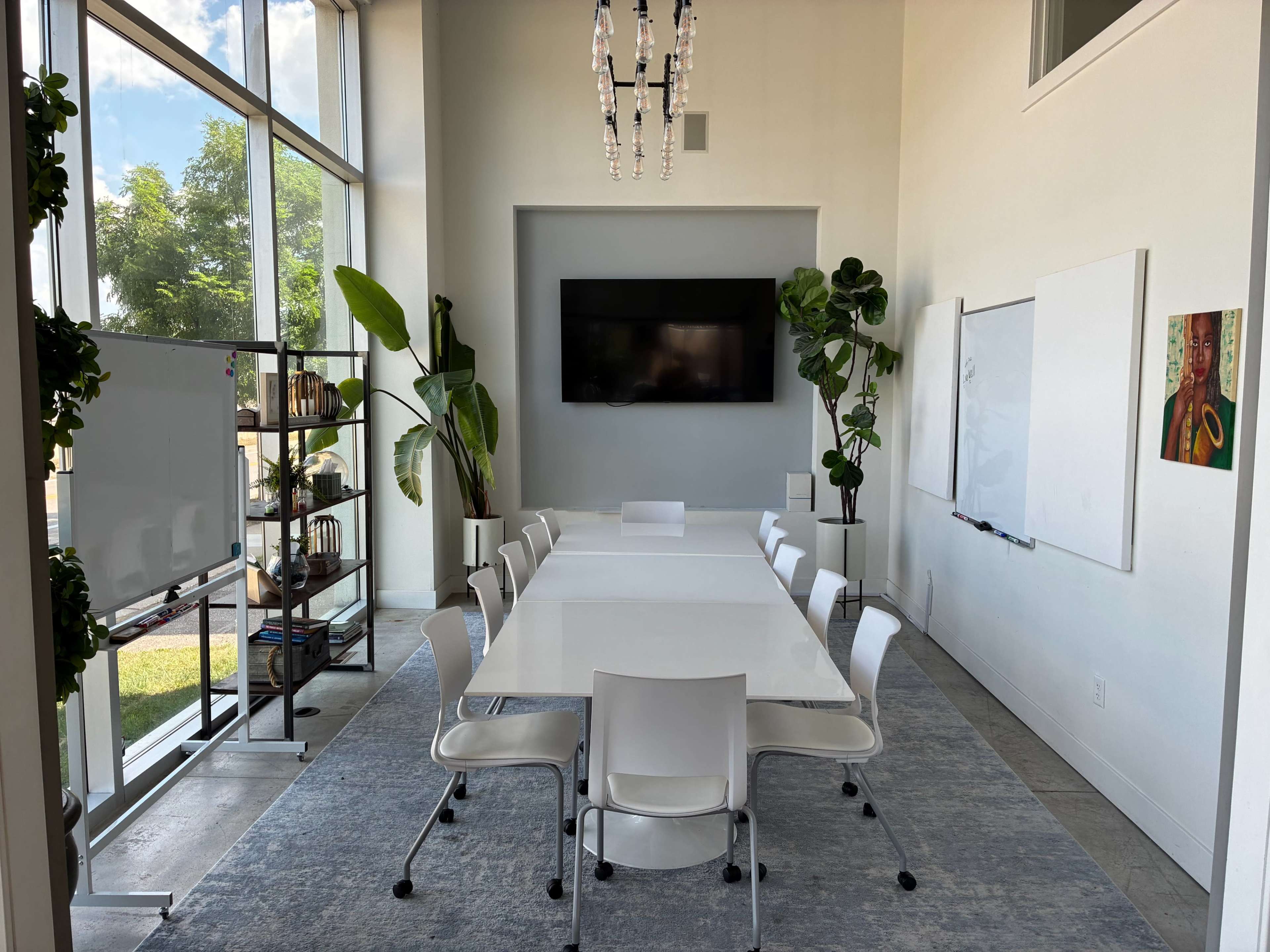 The image shows a modern conference room featuring a long white table surrounded by eight chairs, large windows with views of greenery, and a wall-mounted TV.