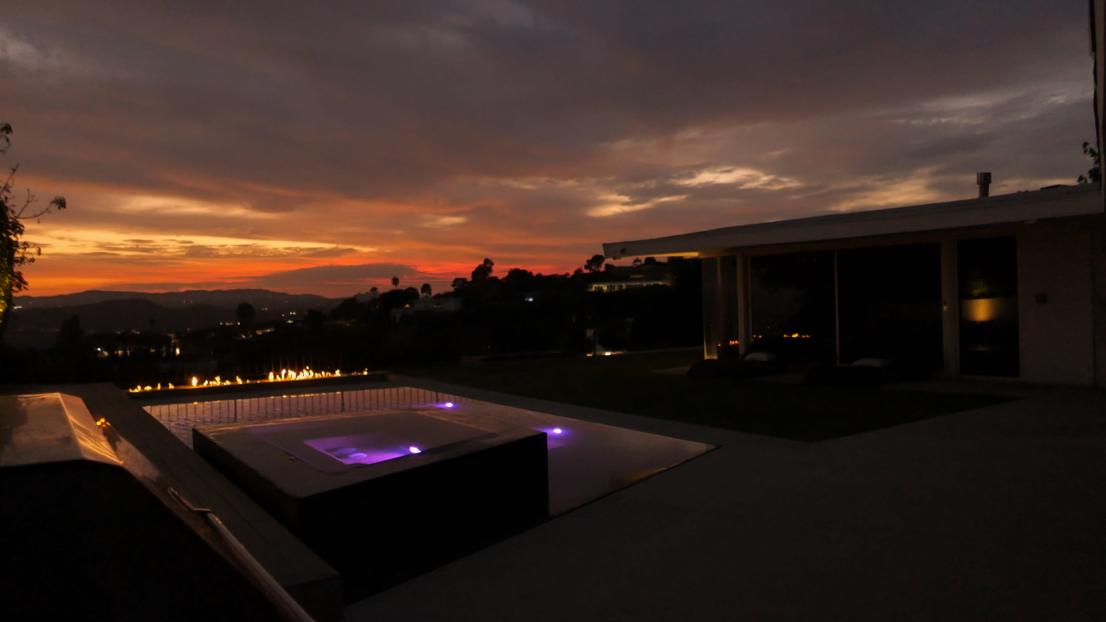 A modern outdoor space features a pool with illuminated edges and a sunset sky filled with clouds and color.