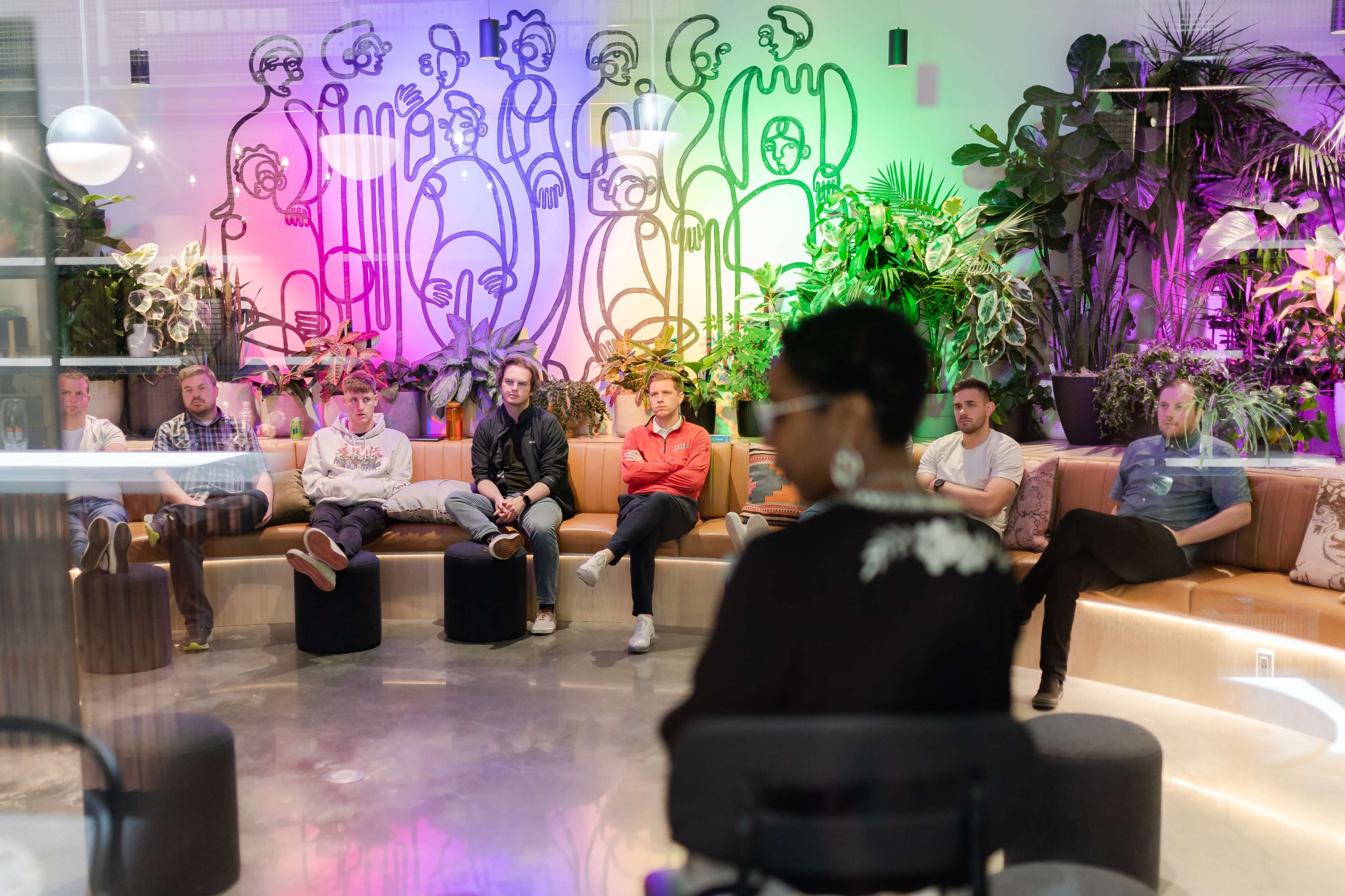 A group of seven people sits in a semi-circle on a modern seating area, while a woman stands in front of them, all in a brightly lit space decorated with colorful wall art and greenery.