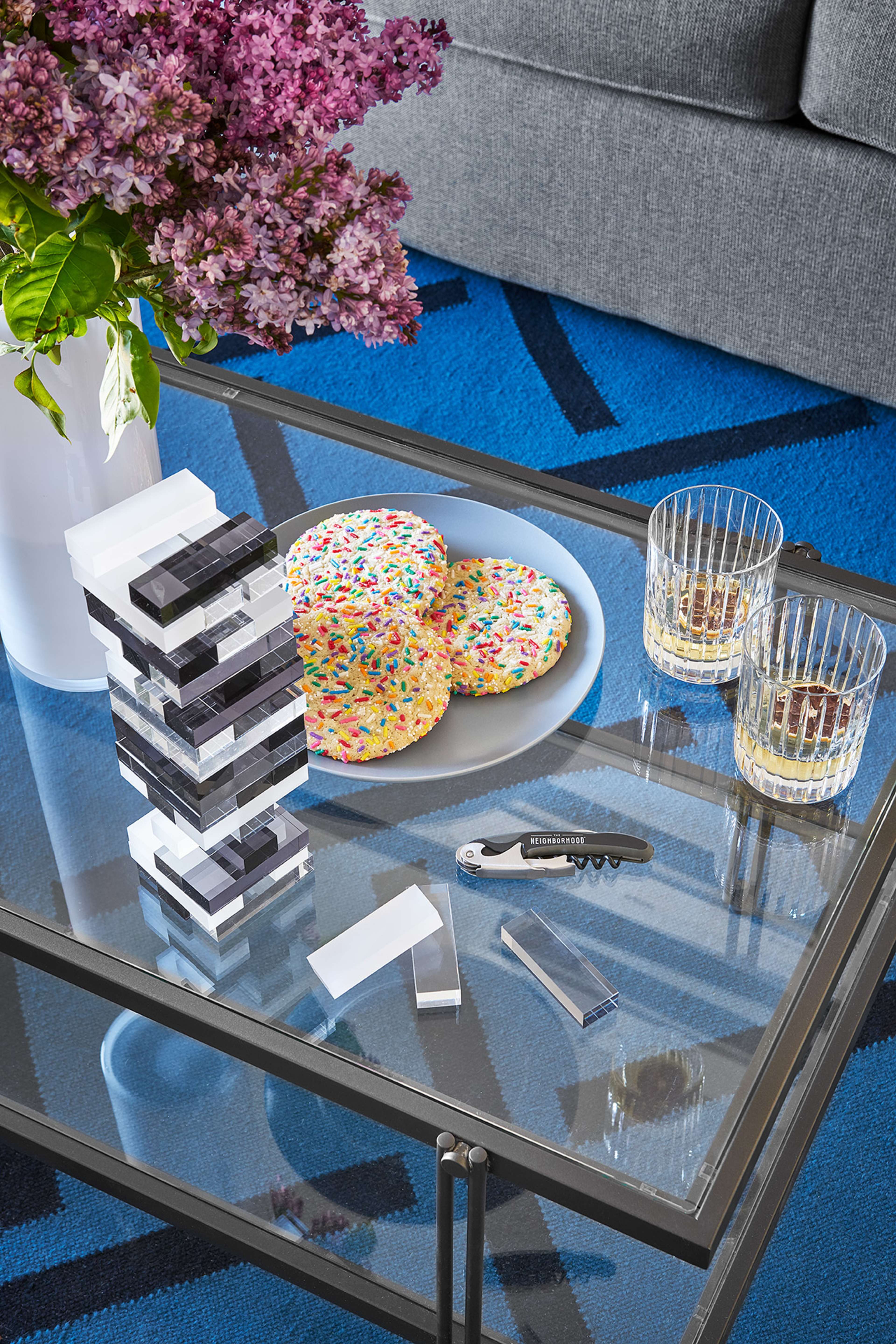 A coffee table features a stack of clear blocks, large colorful cookies on a plate, two glasses with a drink, a small knife, and a vase of lilacs.