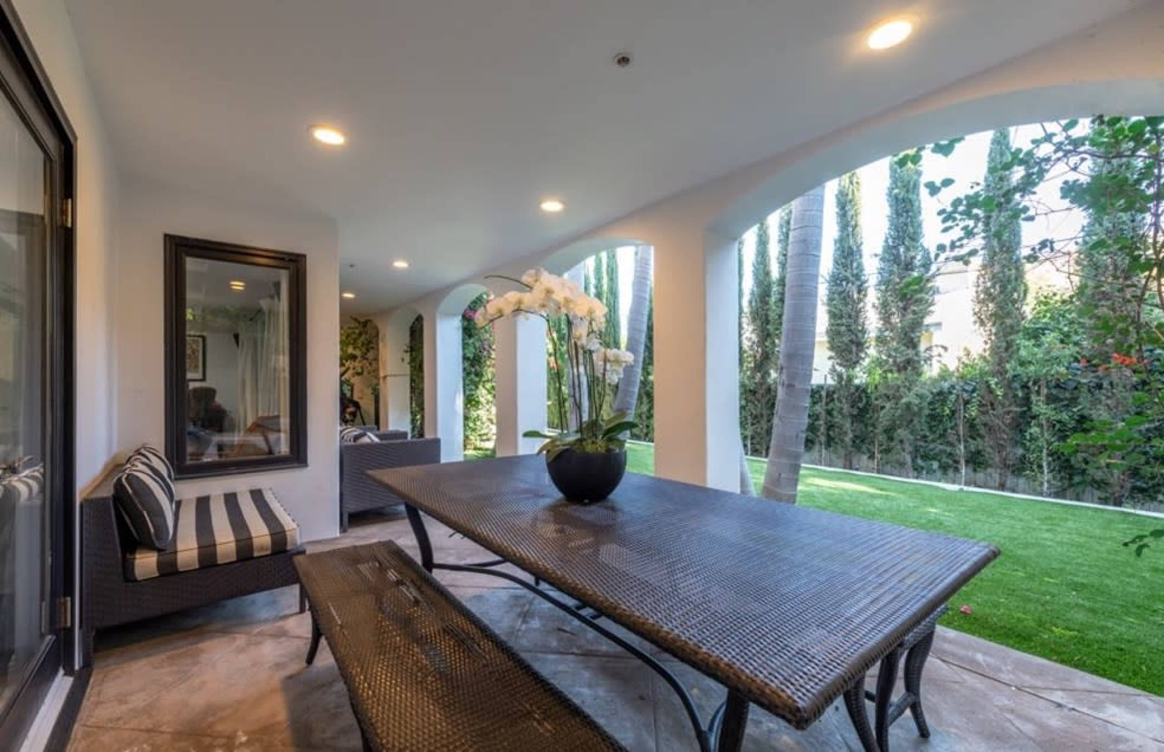 The image shows a covered patio featuring a large dining table and benches, with tall green trees visible in the background.