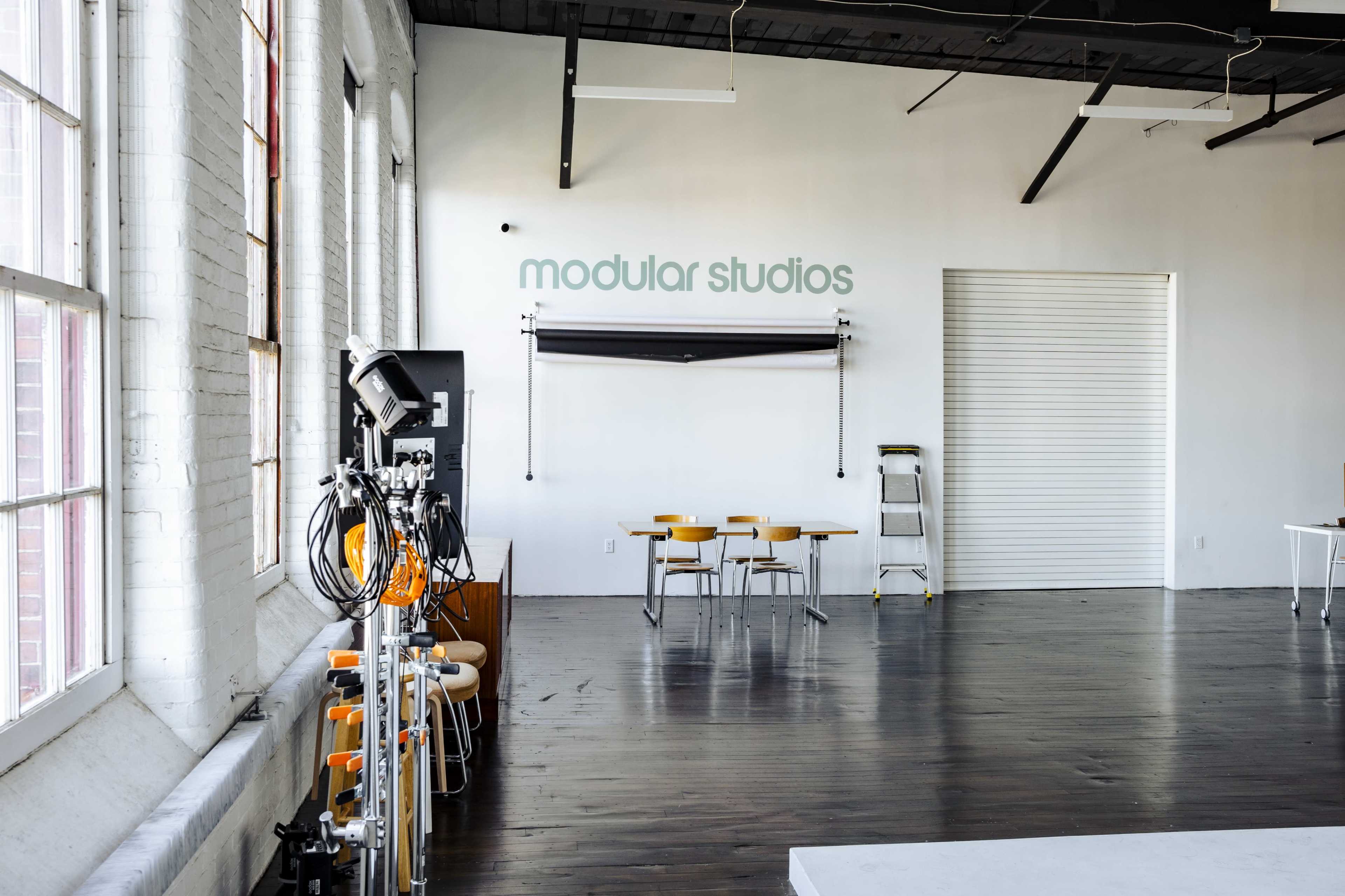 A spacious studio with large windows, a white wall displaying "modular studios," a folding table with chairs, and a rack of lighting equipment.