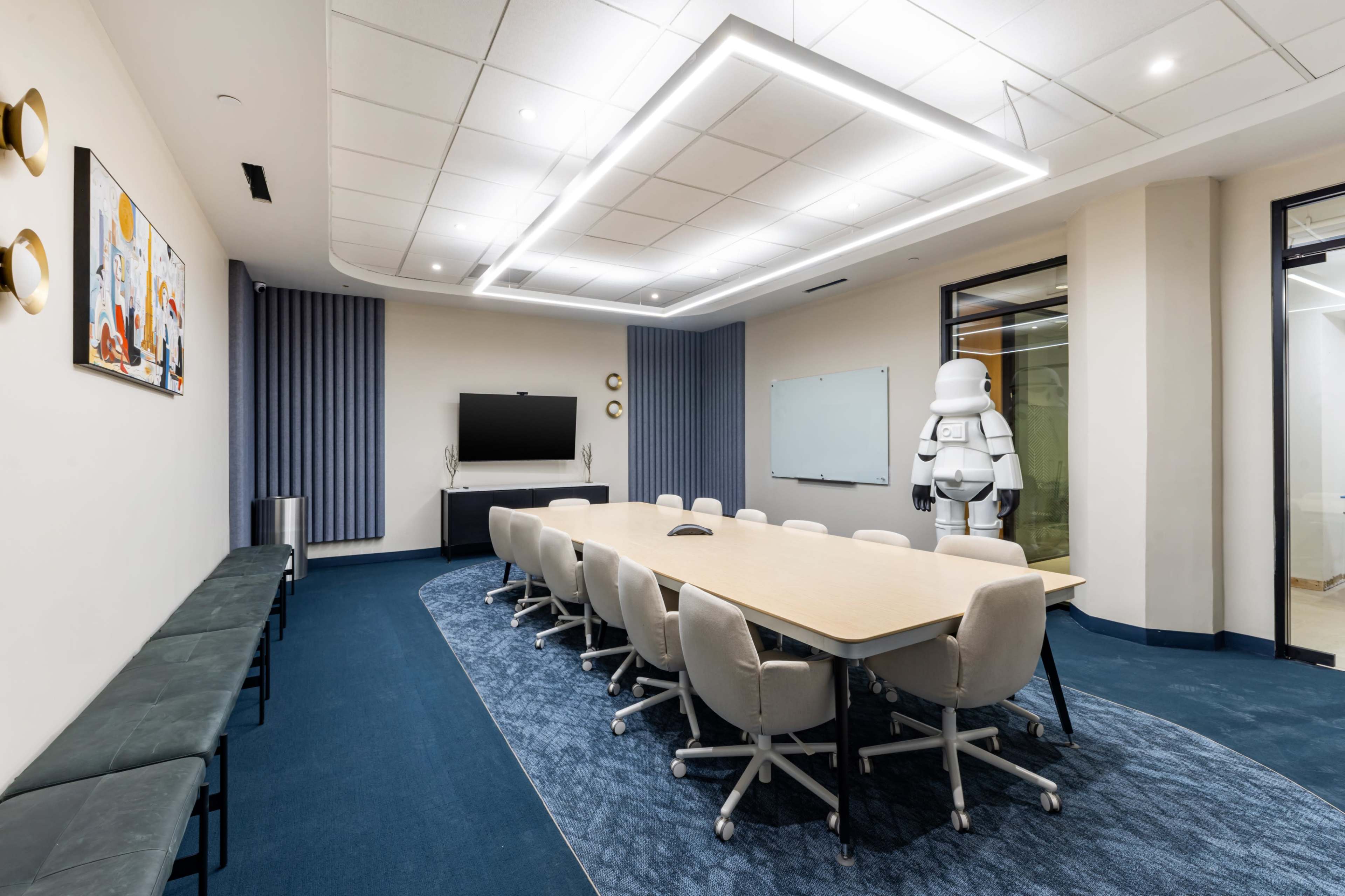 A modern conference room features a large wooden table surrounded by ergonomic chairs, with a TV screen on one wall and a stormtrooper figurine standing beside a whiteboard.