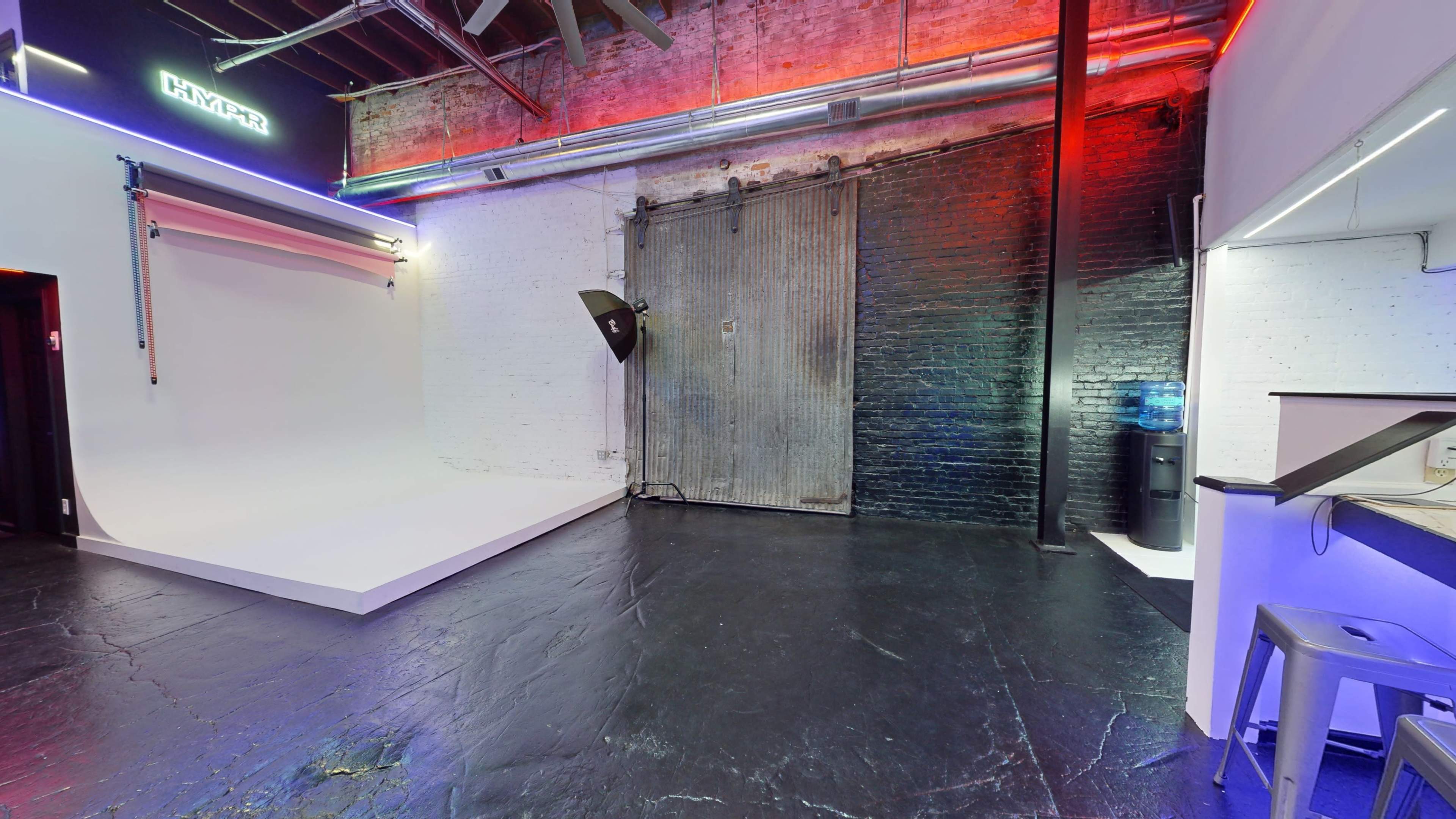A spacious studio with a white backdrop, a corrugated metal door, and a water cooler in the corner, illuminated by colorful lights along the walls.