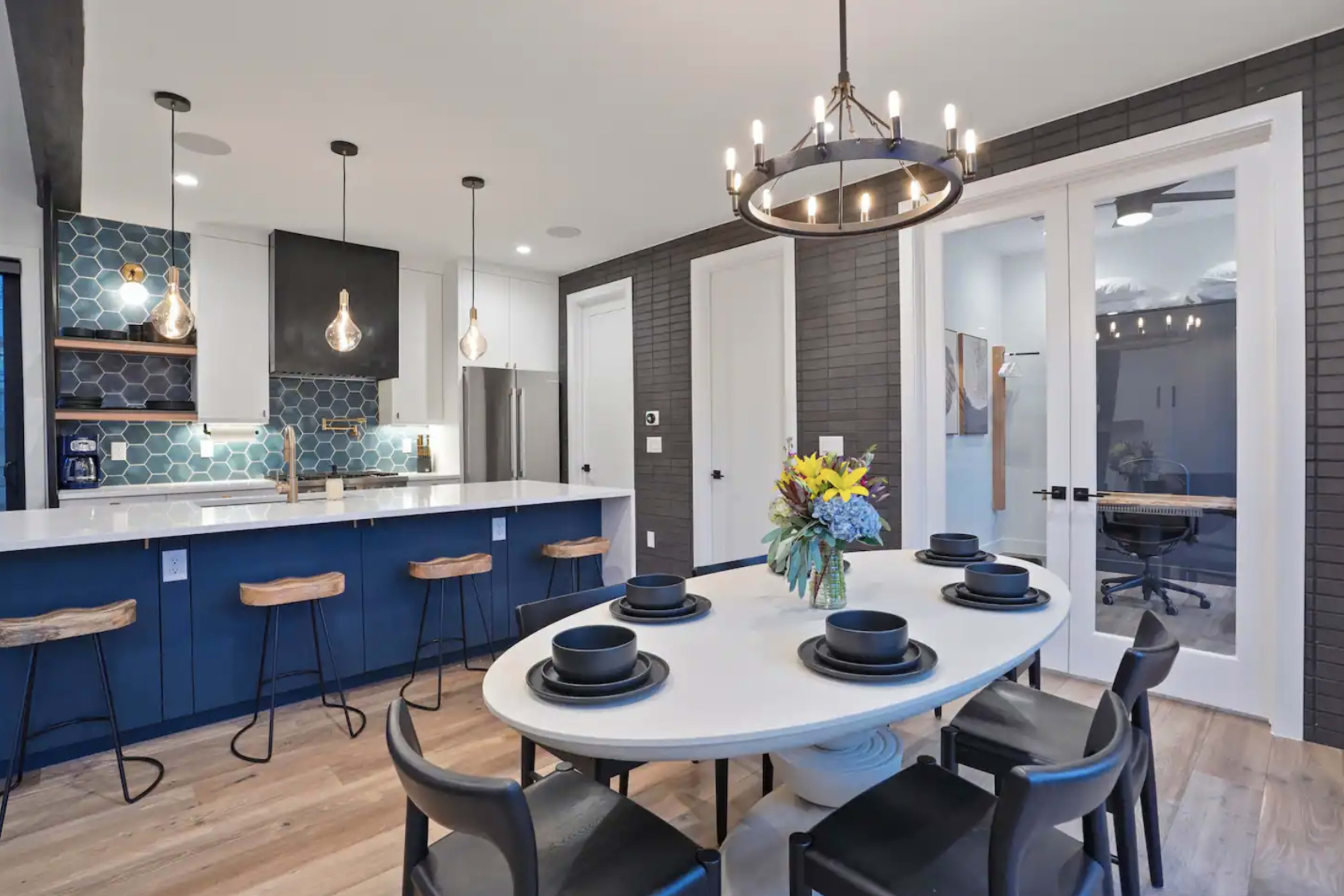 A modern kitchen features a round dining table set for six, with bar stools at the countertop and pendant lights above the island.