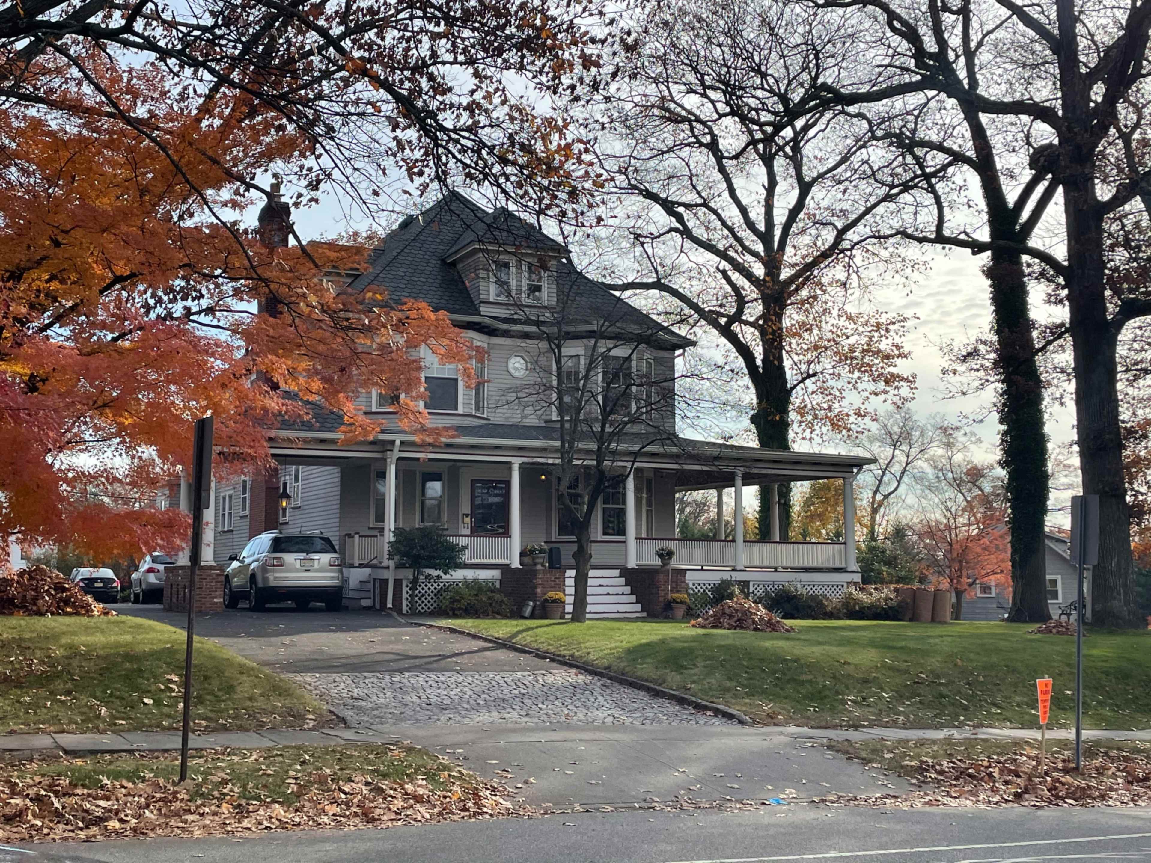 A three-story house with a wrap-around porch is set among autumn trees, displaying orange and yellow foliage.