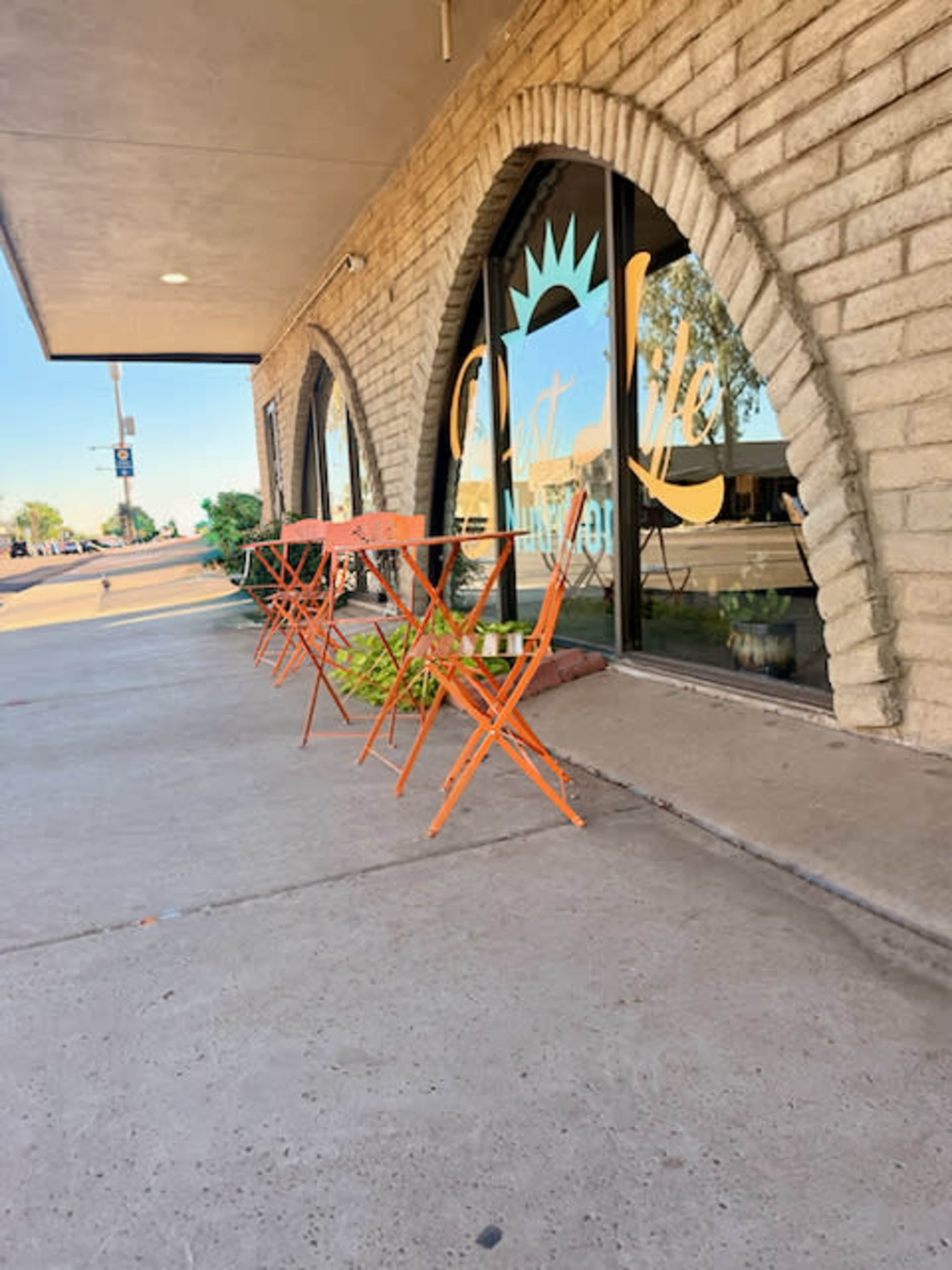 The image shows a sidewalk cafe with orange metal chairs and tables positioned outside a building with arched windows.