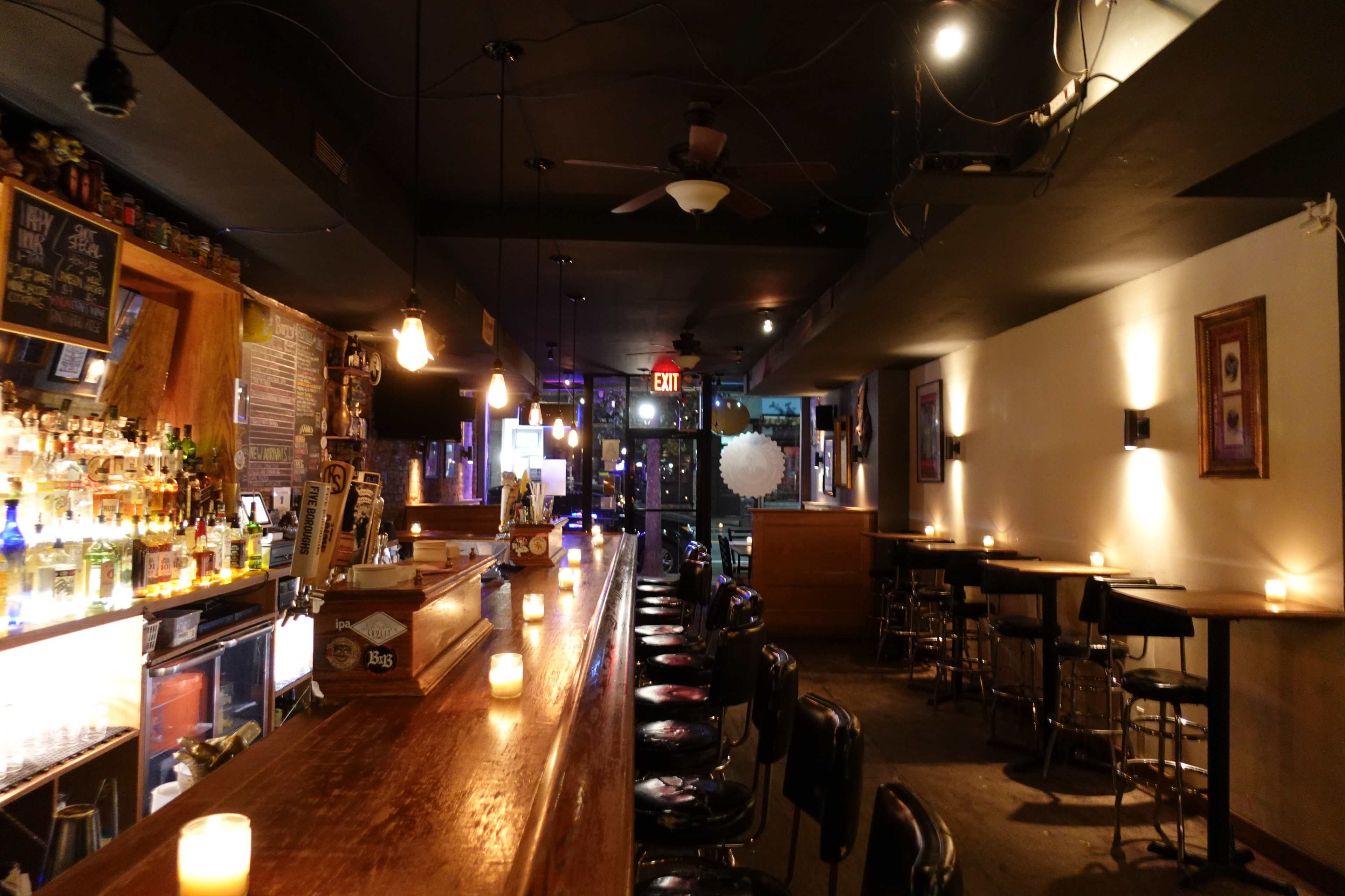 The interior of a bar with a long wooden counter, shelves stocked with bottles behind the bar, and tables with candles arranged throughout the space.