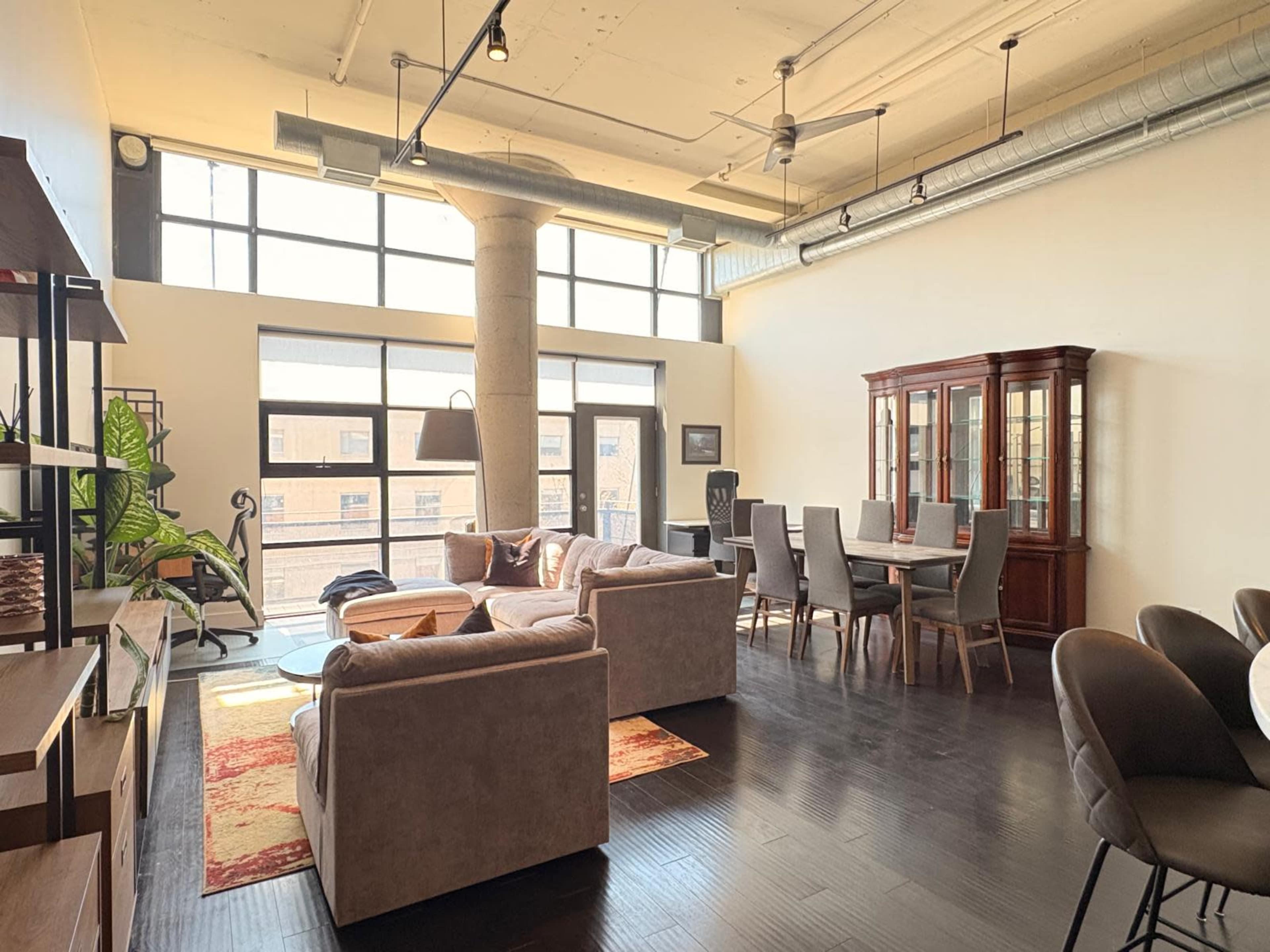 The image depicts a spacious, modern living area featuring a cozy seating arrangement, a dining table, and large windows allowing natural light to fill the space.