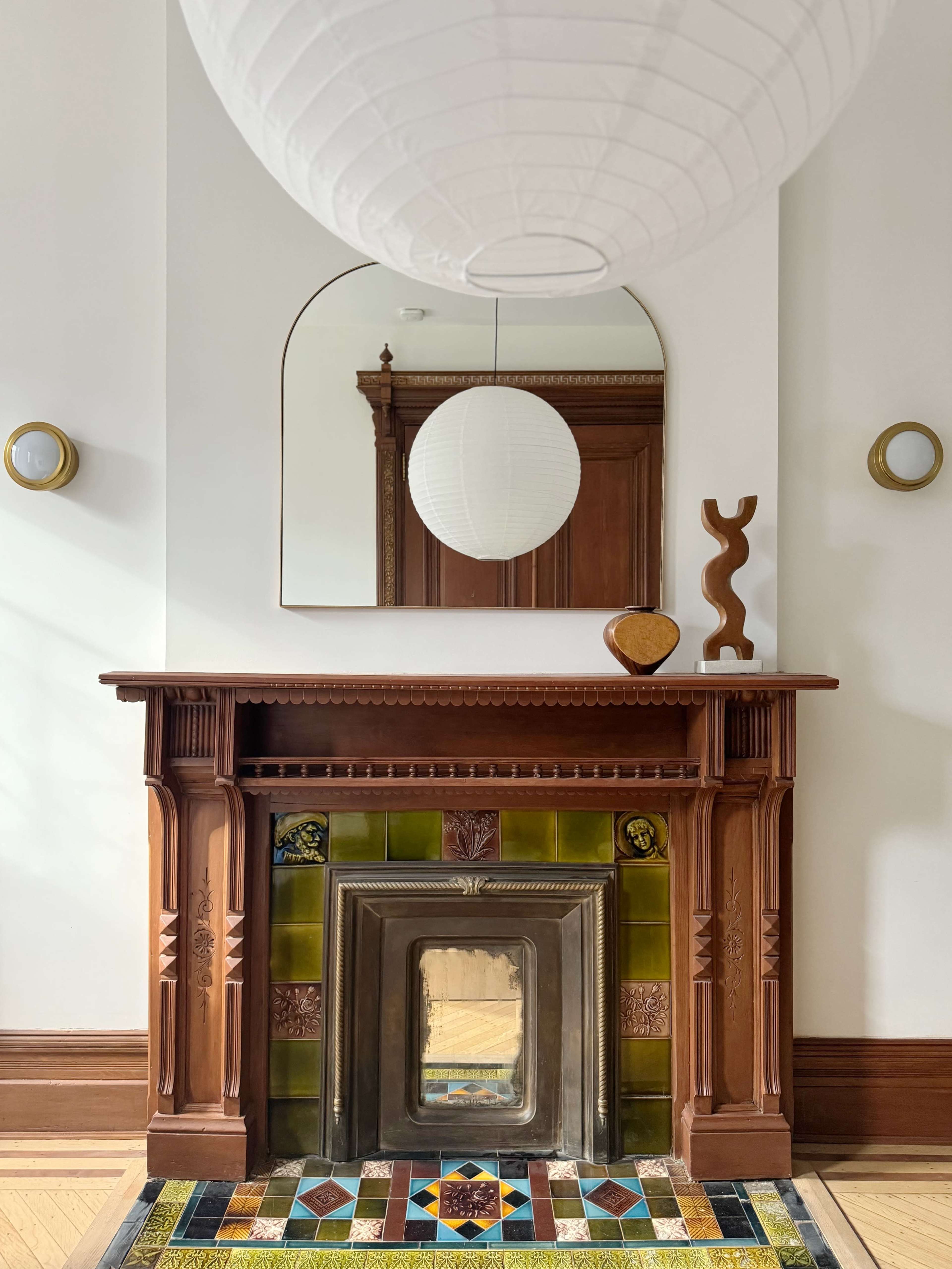 The image shows a decorative fireplace with intricate tiles and a large mirror above it, complemented by a round pendant light and a wooden sculpture on the mantel.
