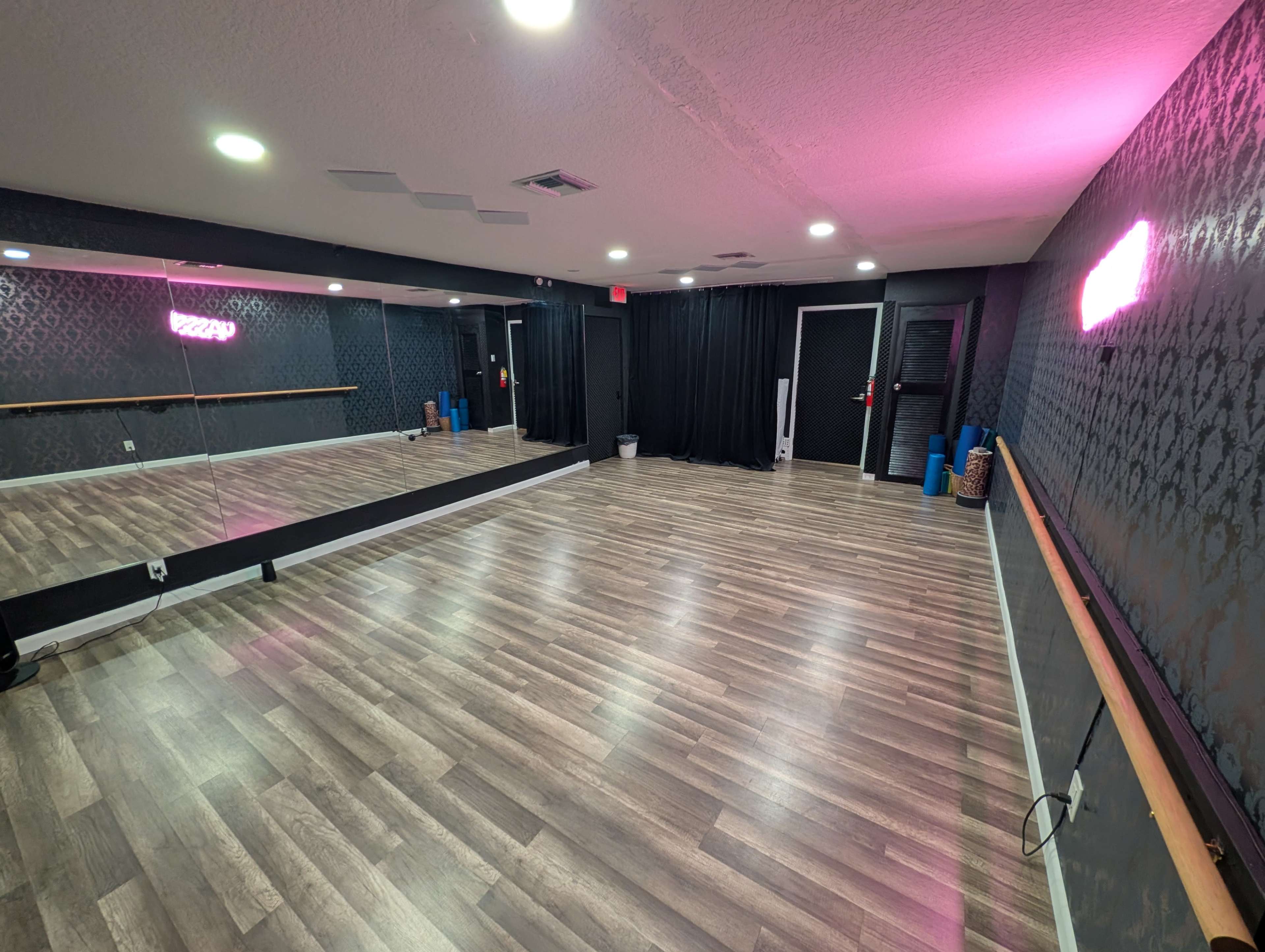 The image shows a spacious dance studio with wooden flooring, mirrors on one wall, and soft pink lighting.